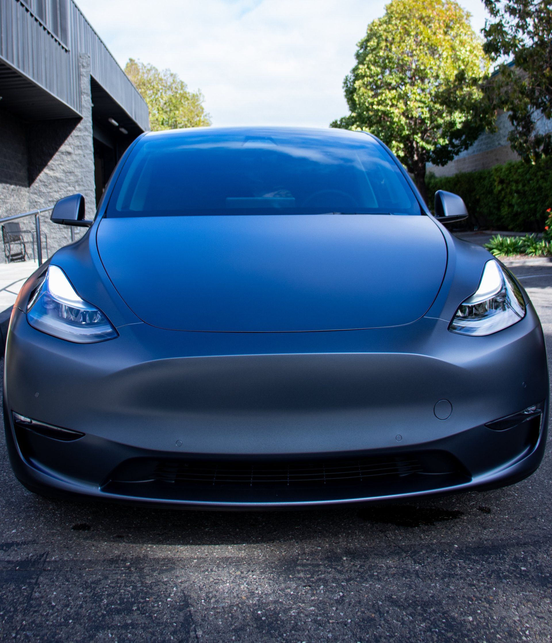 Front view of a dark gray Tesla parked outdoors in a driveway, with a building and trees in the background.