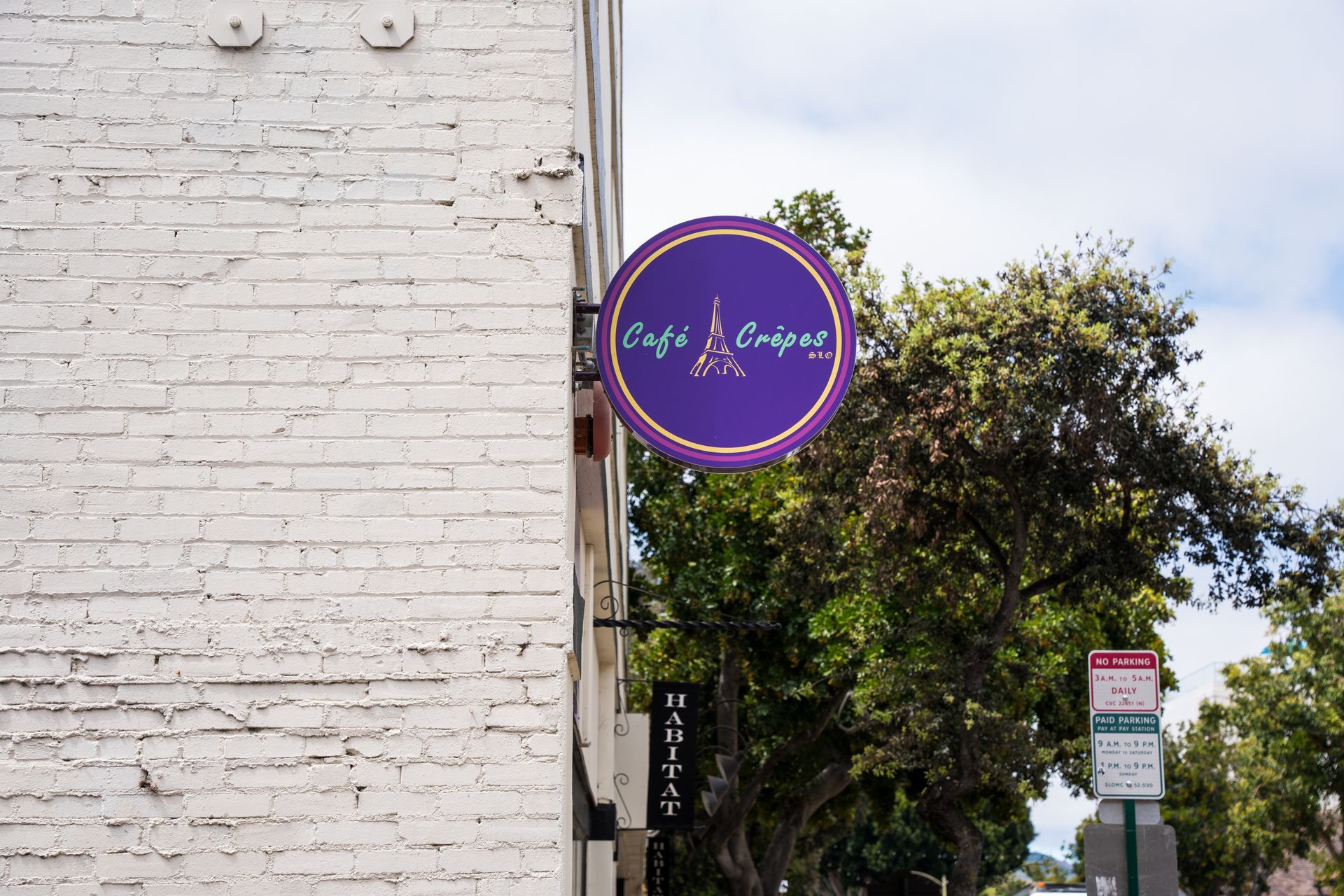 Purple circular shop sign on a white brick wall beside a tree-lined street.