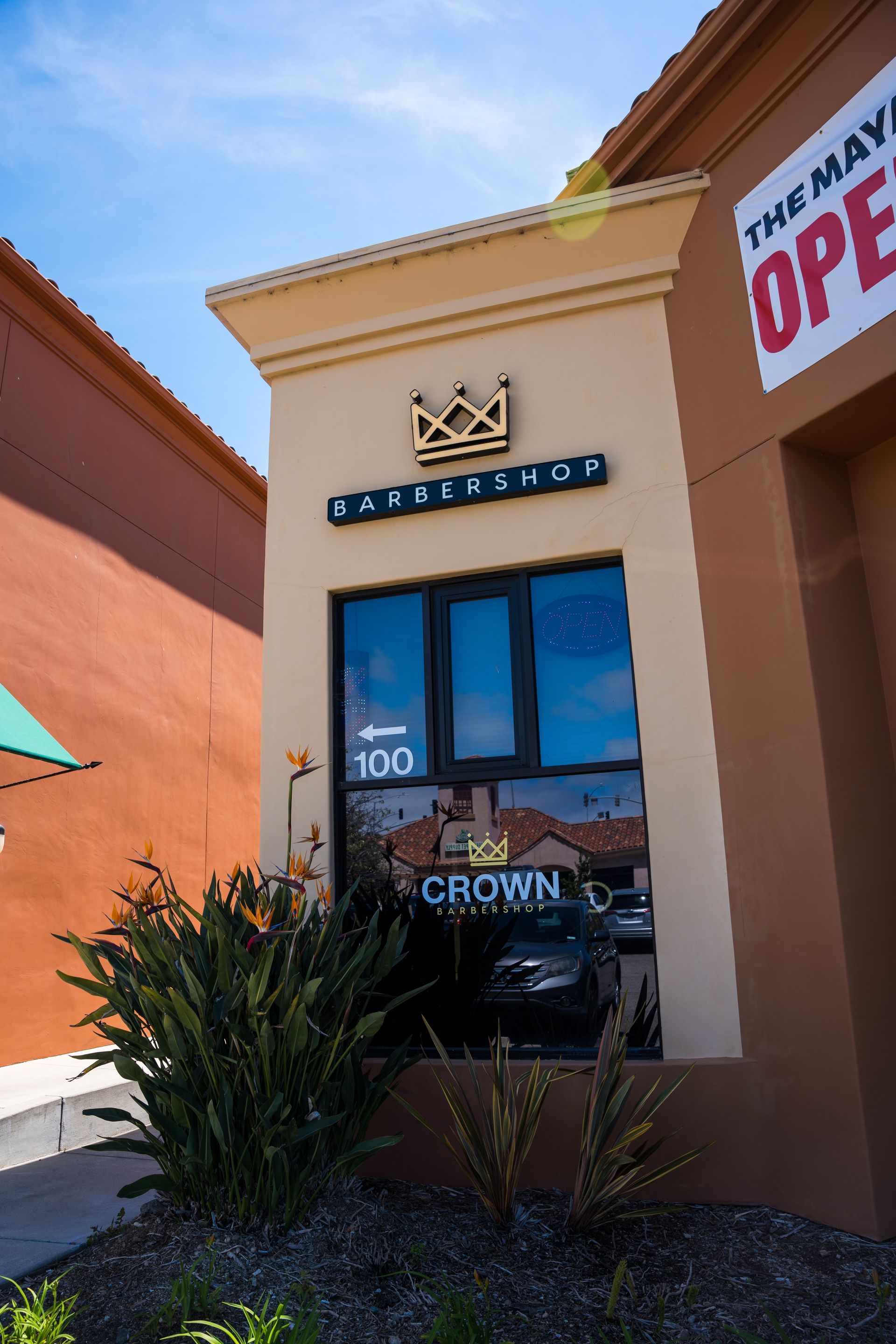 Storefront entrance with blue signage, tan stucco walls, and a “Now Open” banner.