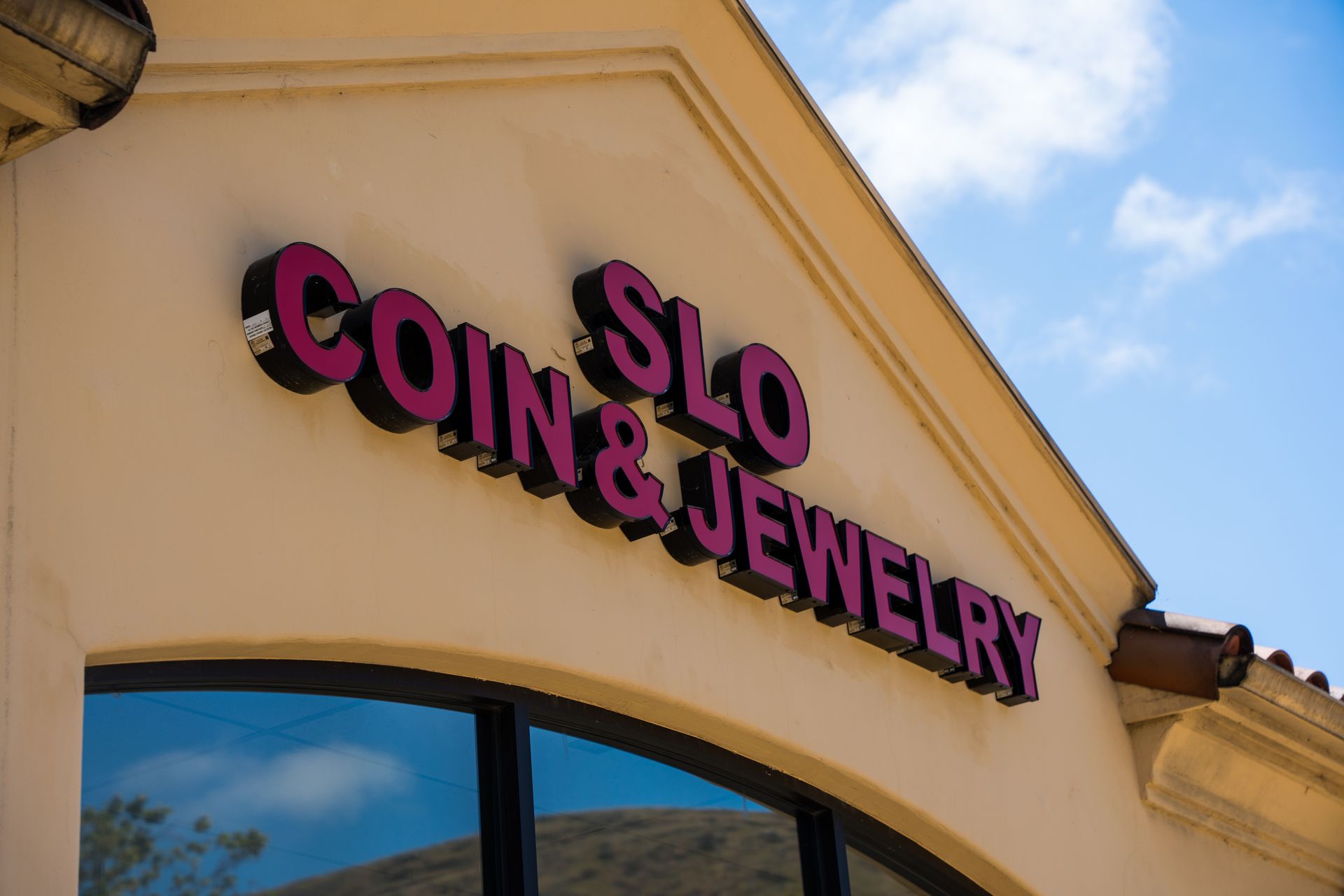 SLO Coin & Jewelry storefront sign in pink letters on a beige building under a blue sky