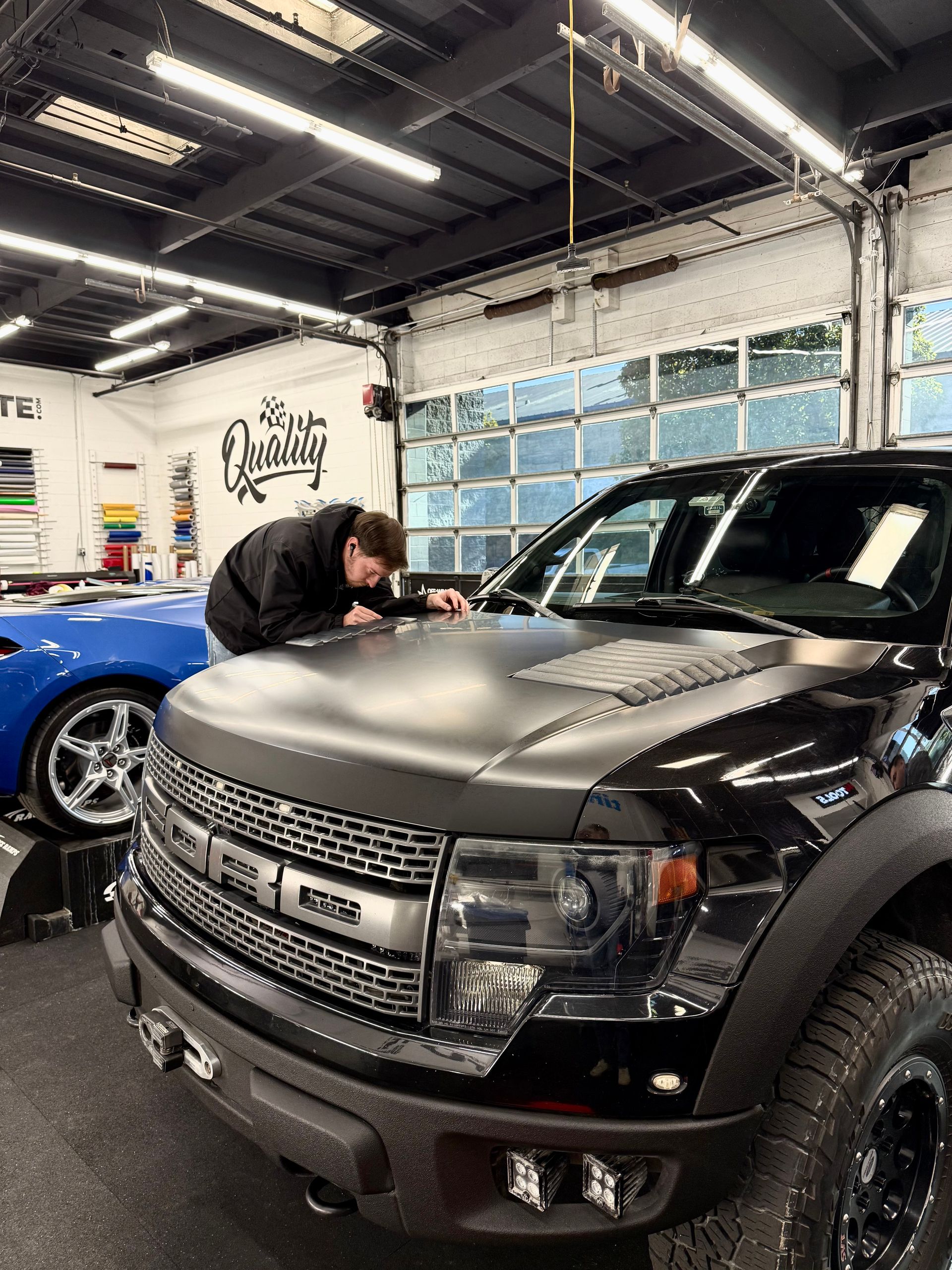 Person leaning over the hood of a black Ford truck in a garage, with a blue car beside it.