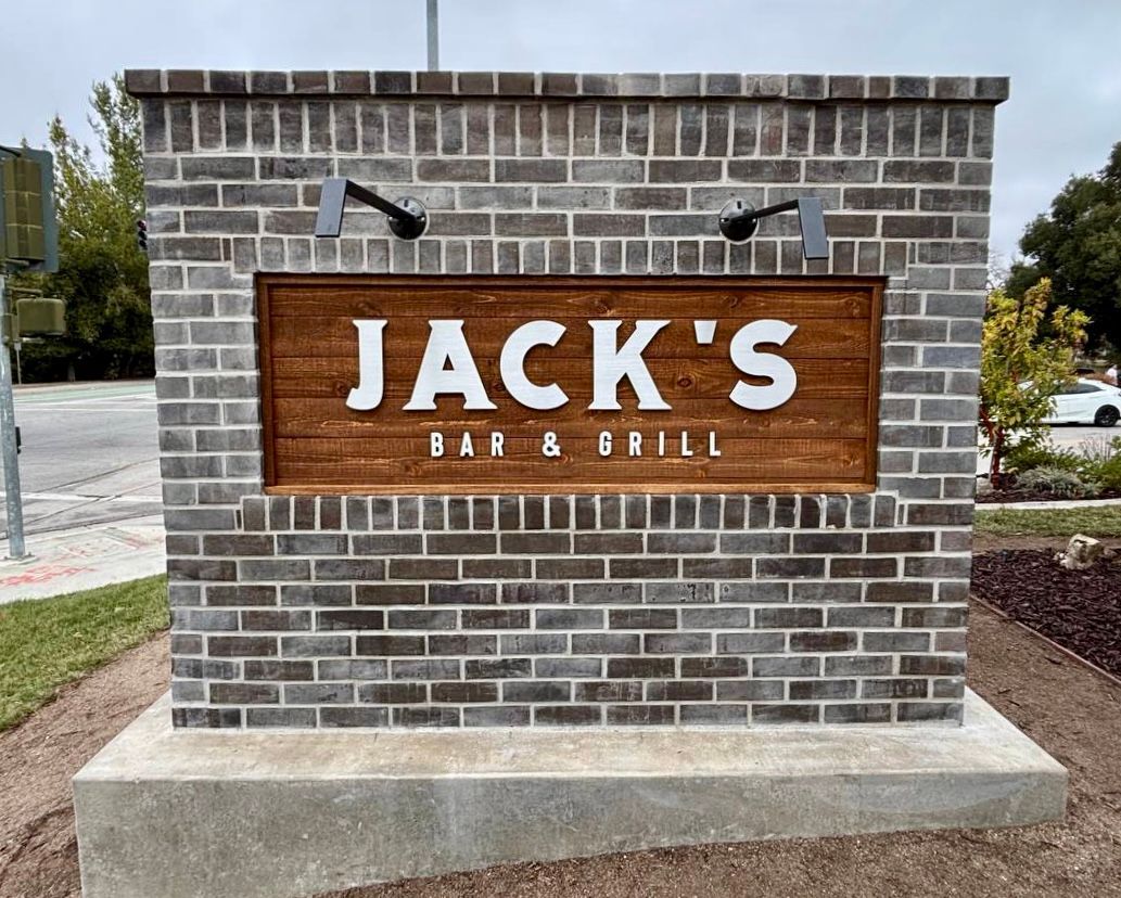 Brick monument sign for Jack’s Bar & Grill with wood panel and black lettering outside.