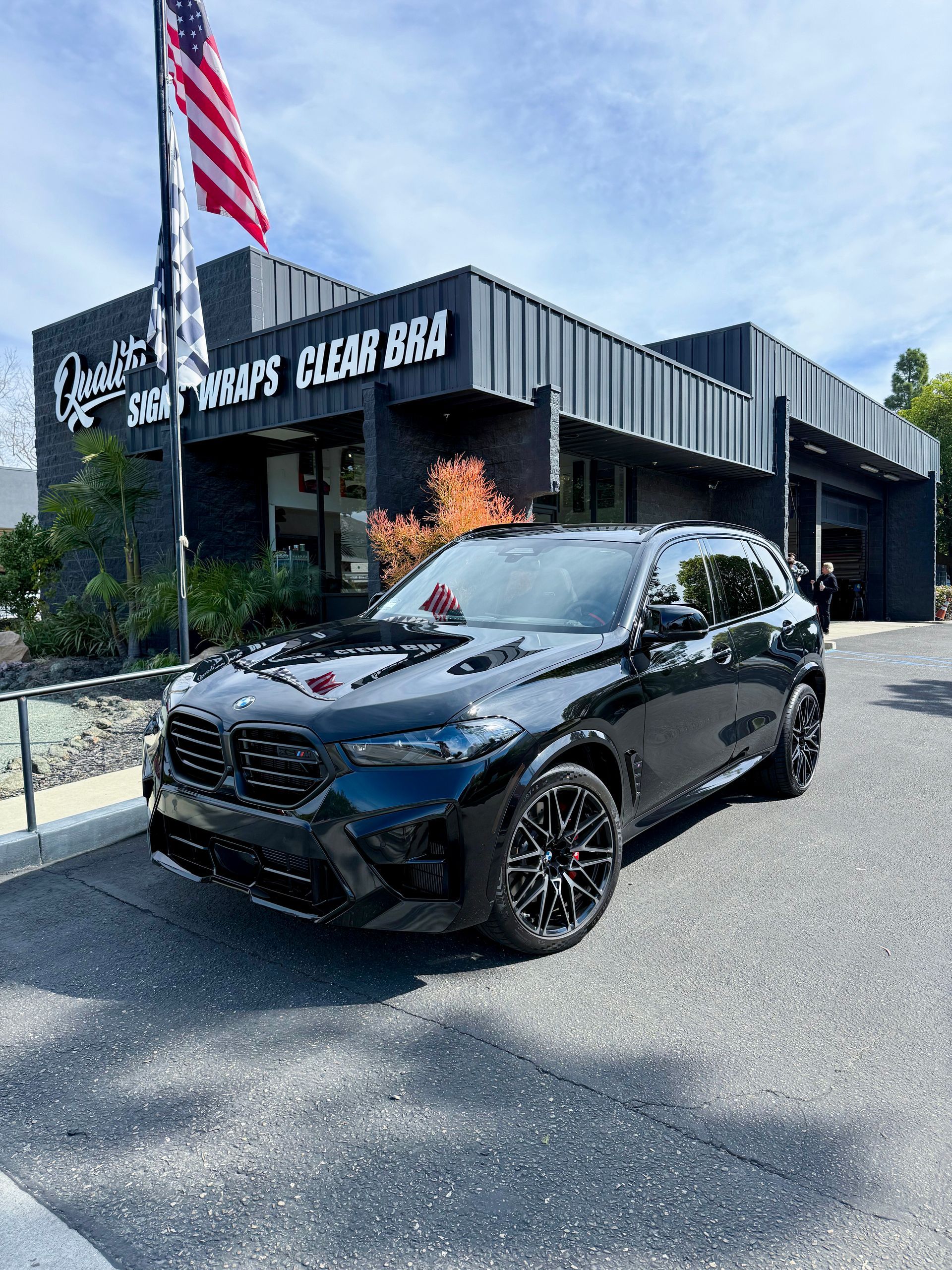 Black BMW SUV parked outside a race shop with an American flag and sunny shadows