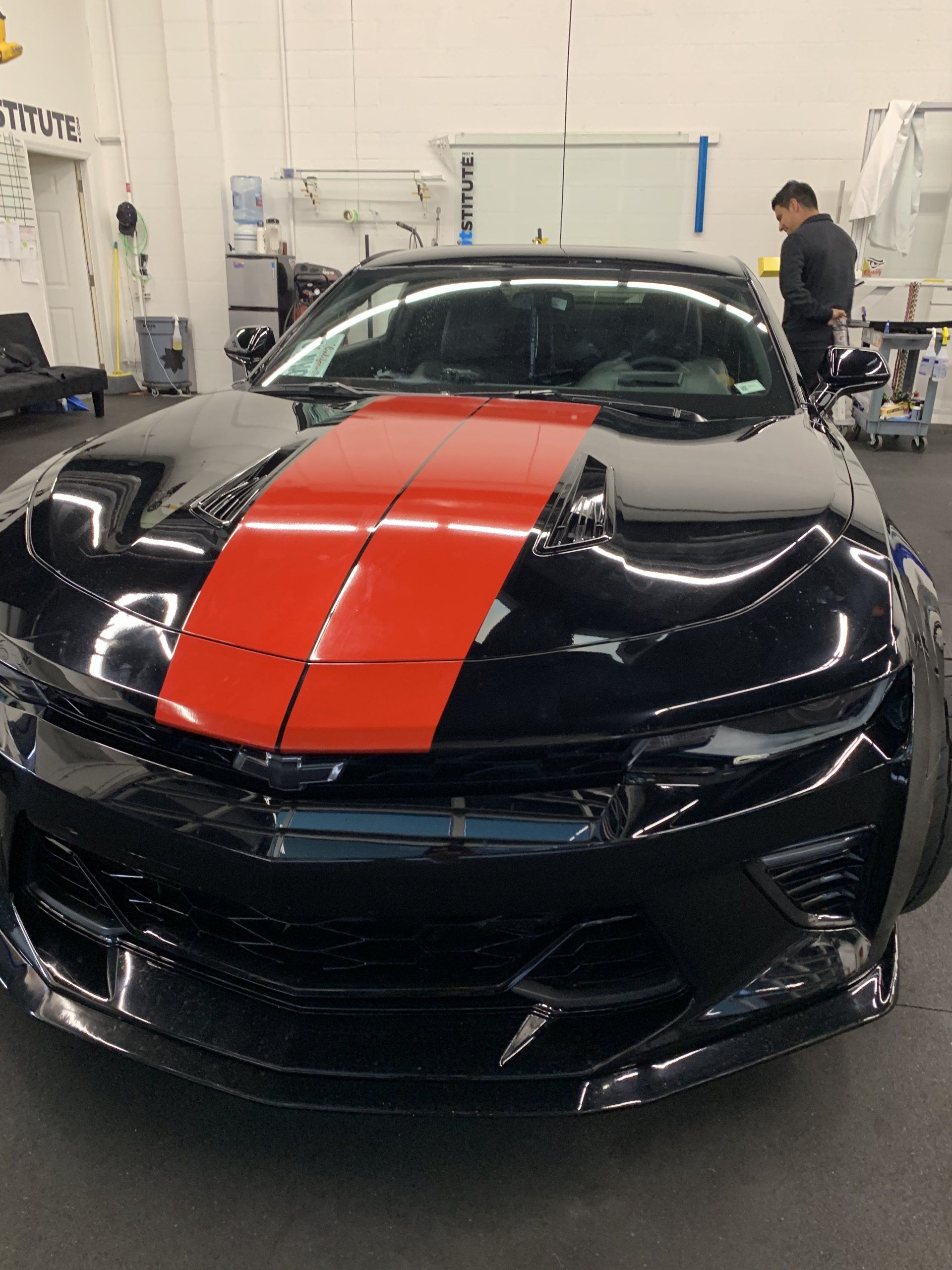 Black sports car with a red racing stripe in a workshop, with a person working in the background