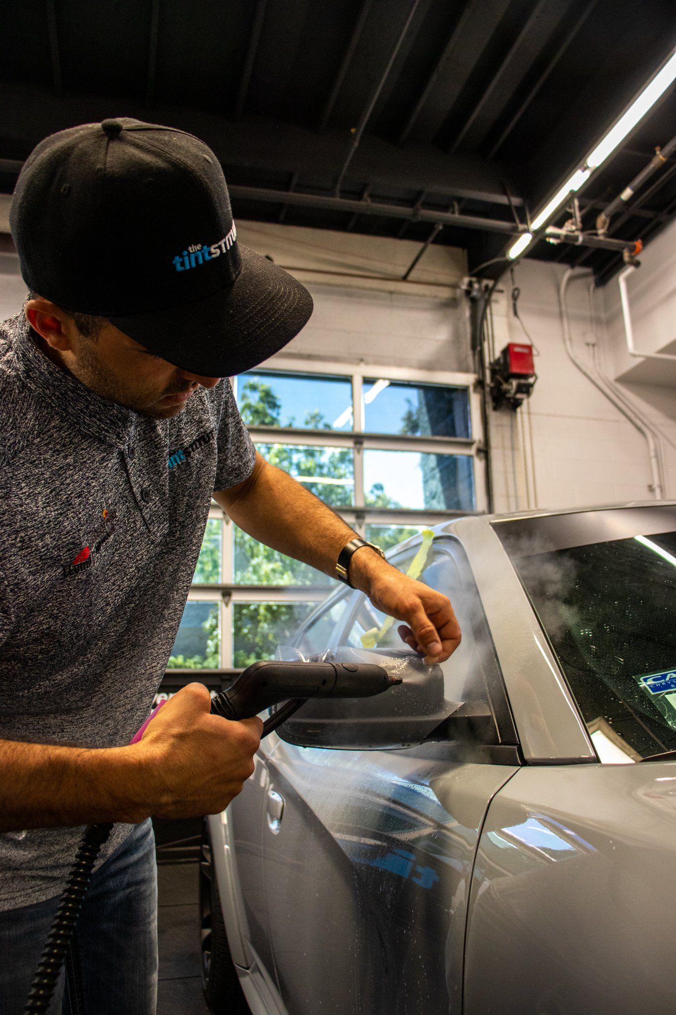 Man polishing a gray car in a garage, working near the windshield with a cloth