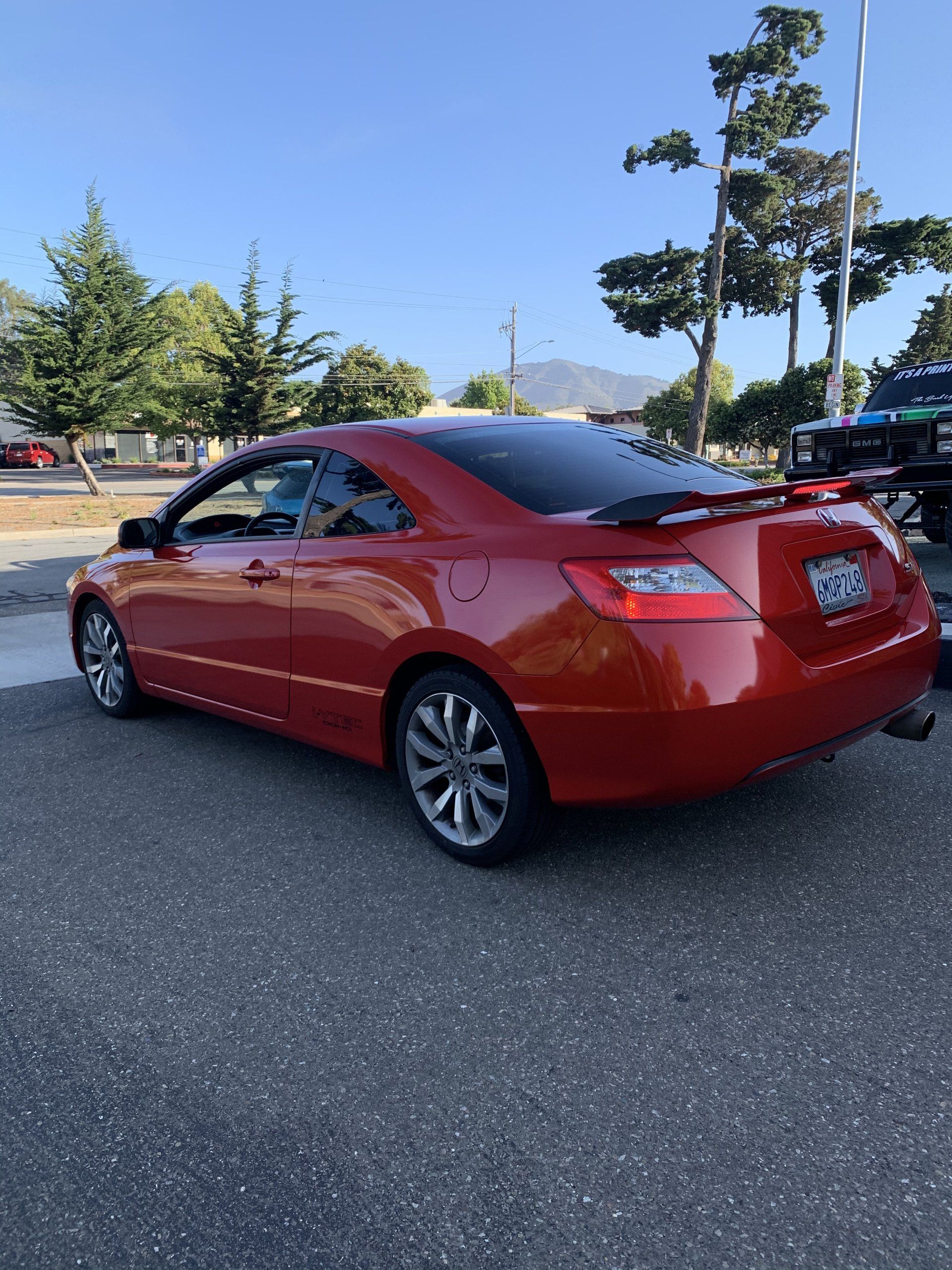 Red coupe parked in a lot on a sunny day, with trees and mountains in the background.