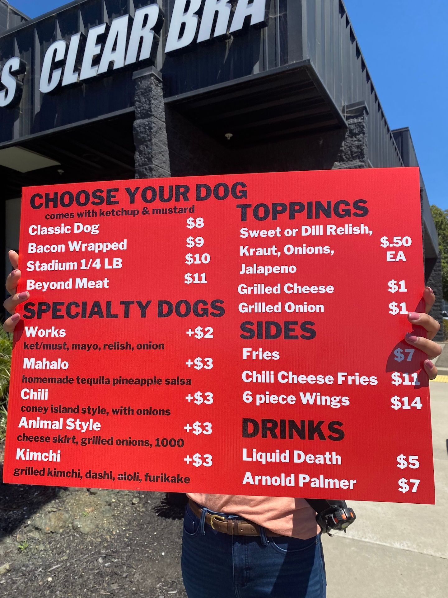 Red hot dog stand menu board with specials, toppings, sides, and drinks under a sign reading Clear B