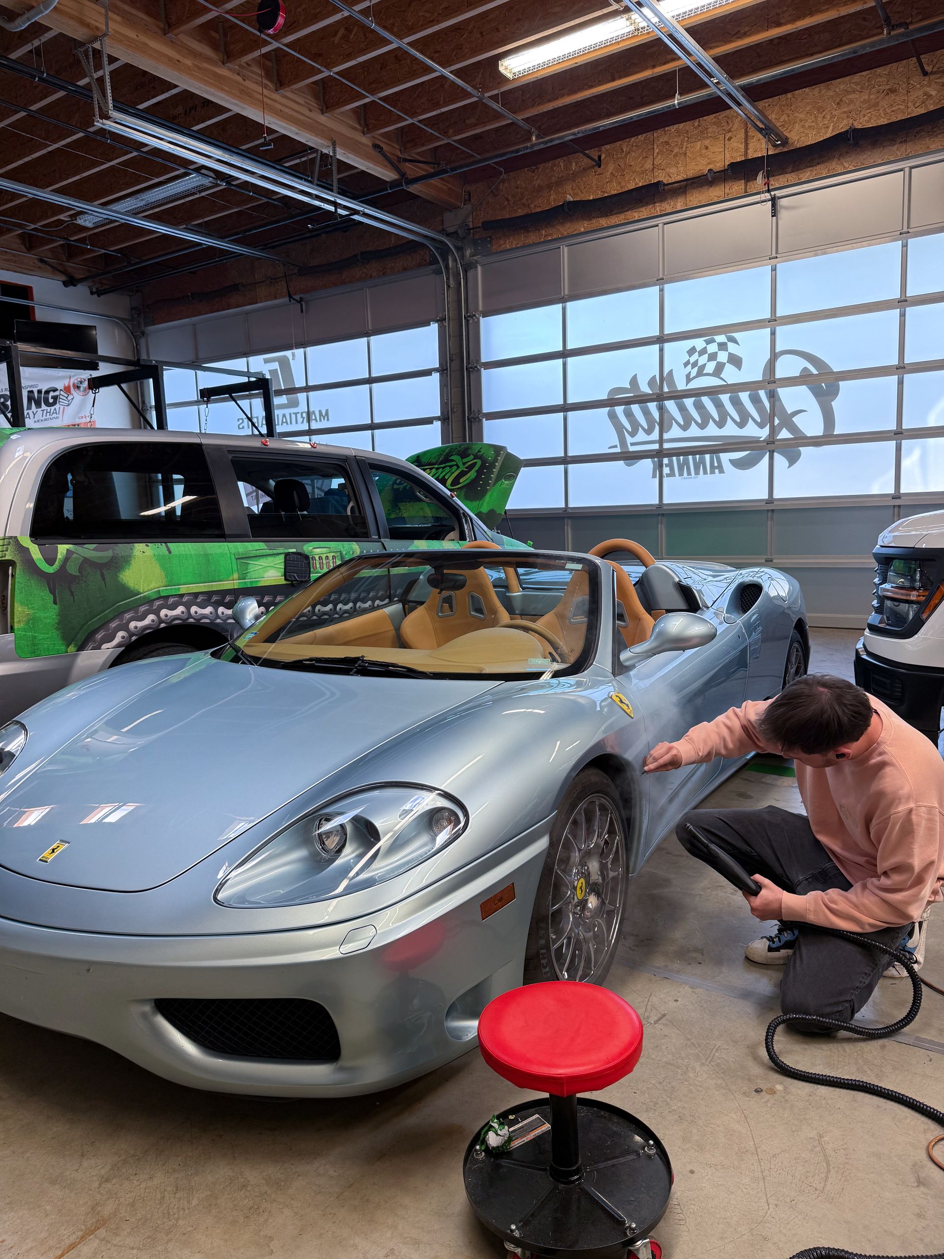 Silver sports car in a garage, with a man crouching beside it and a green van behind.