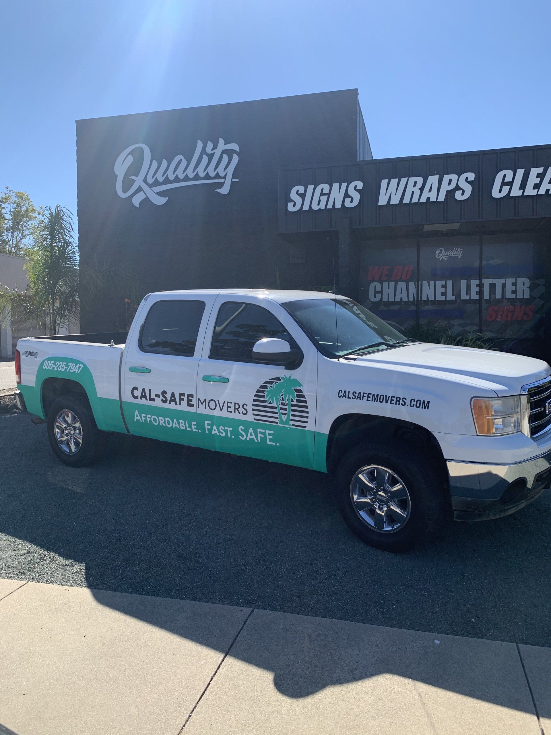 White and green utility truck parked in front of a sign shop with “Quality” and “Signs Wraps” on the building