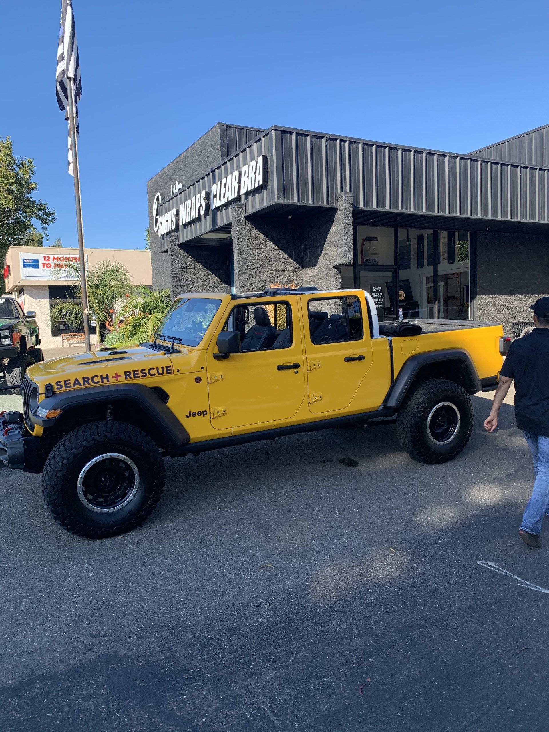 Yellow lifted Jeep parked on a street outside a black storefront, with a person walking nearby