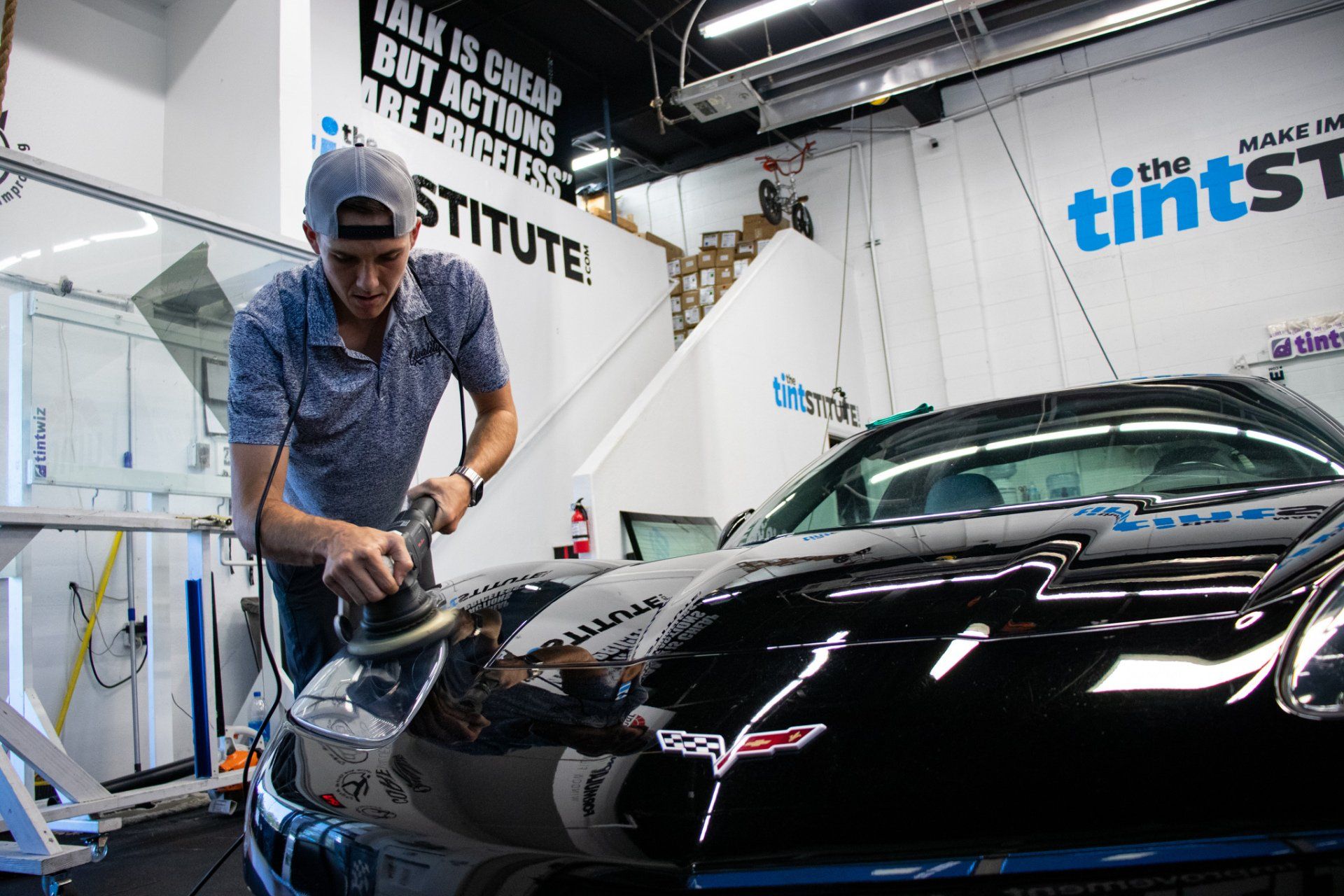 Man polishing a black sports car in a detailing shop with bright lights and tint signs