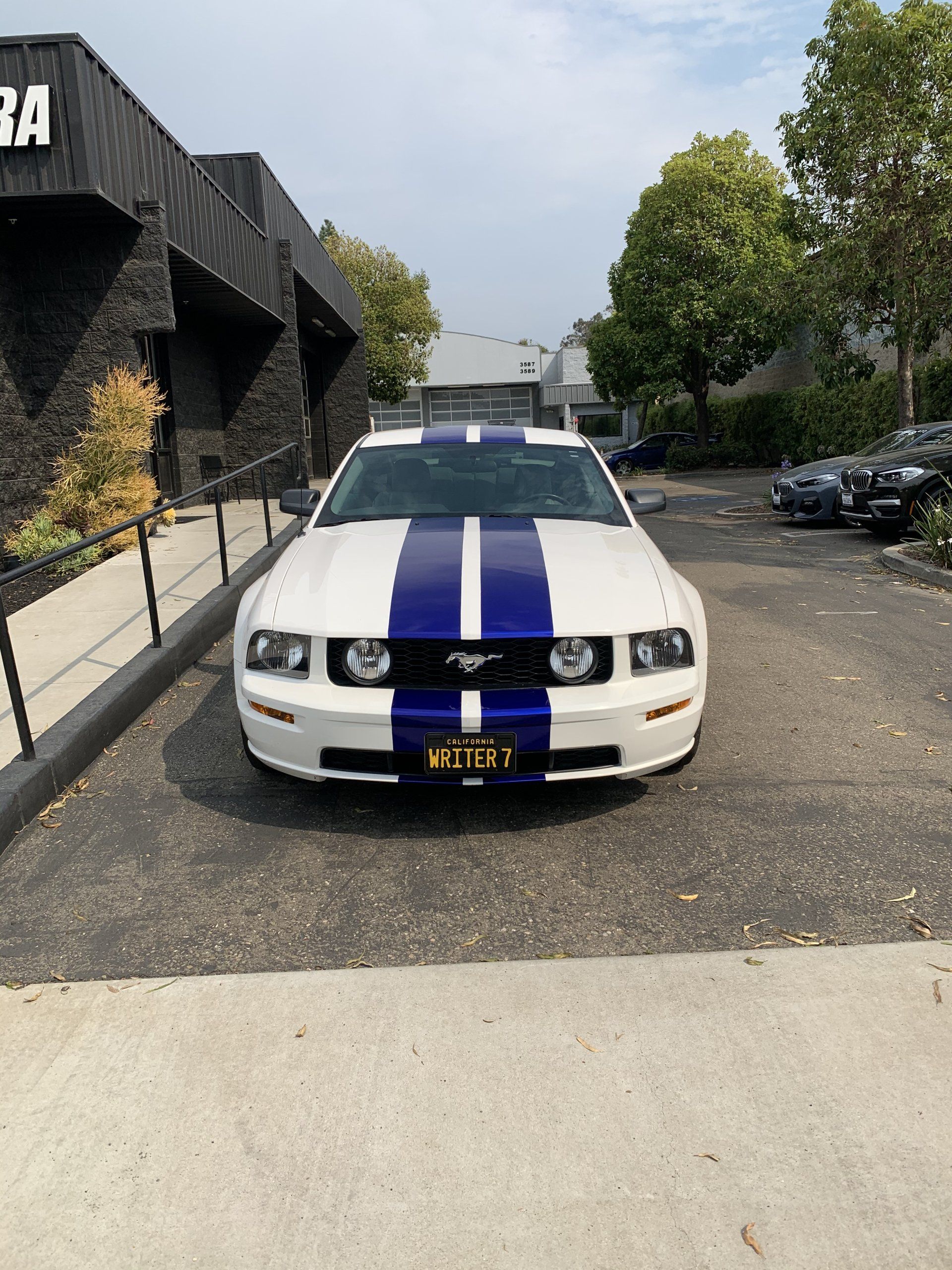 White Mustang with blue racing stripes parked on a gravel lot beside a modern building.