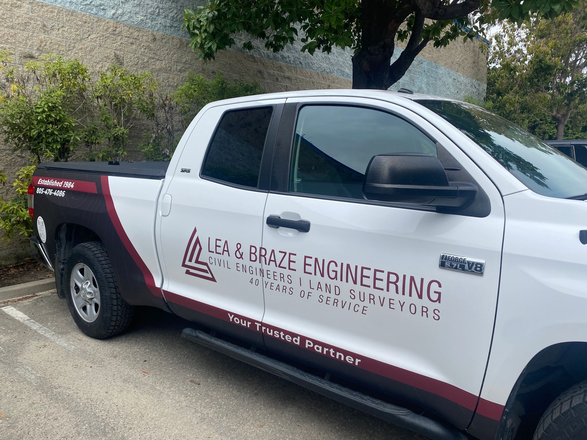 White and black pickup truck with “La & Braz Engineering” logo parked by a roadside wall
