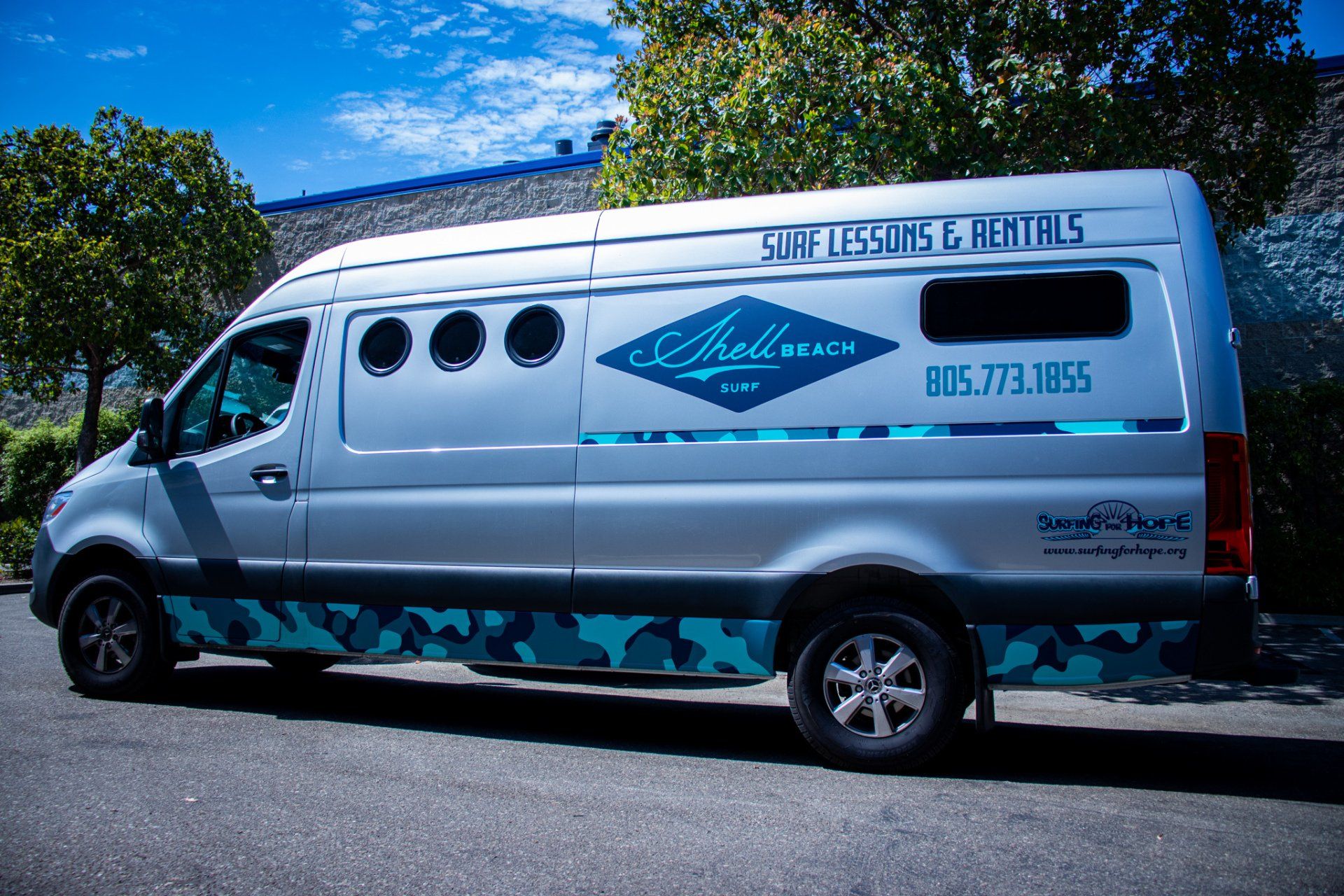 Silver utility van with teal graphics and company logo parked outdoors under a blue sky.