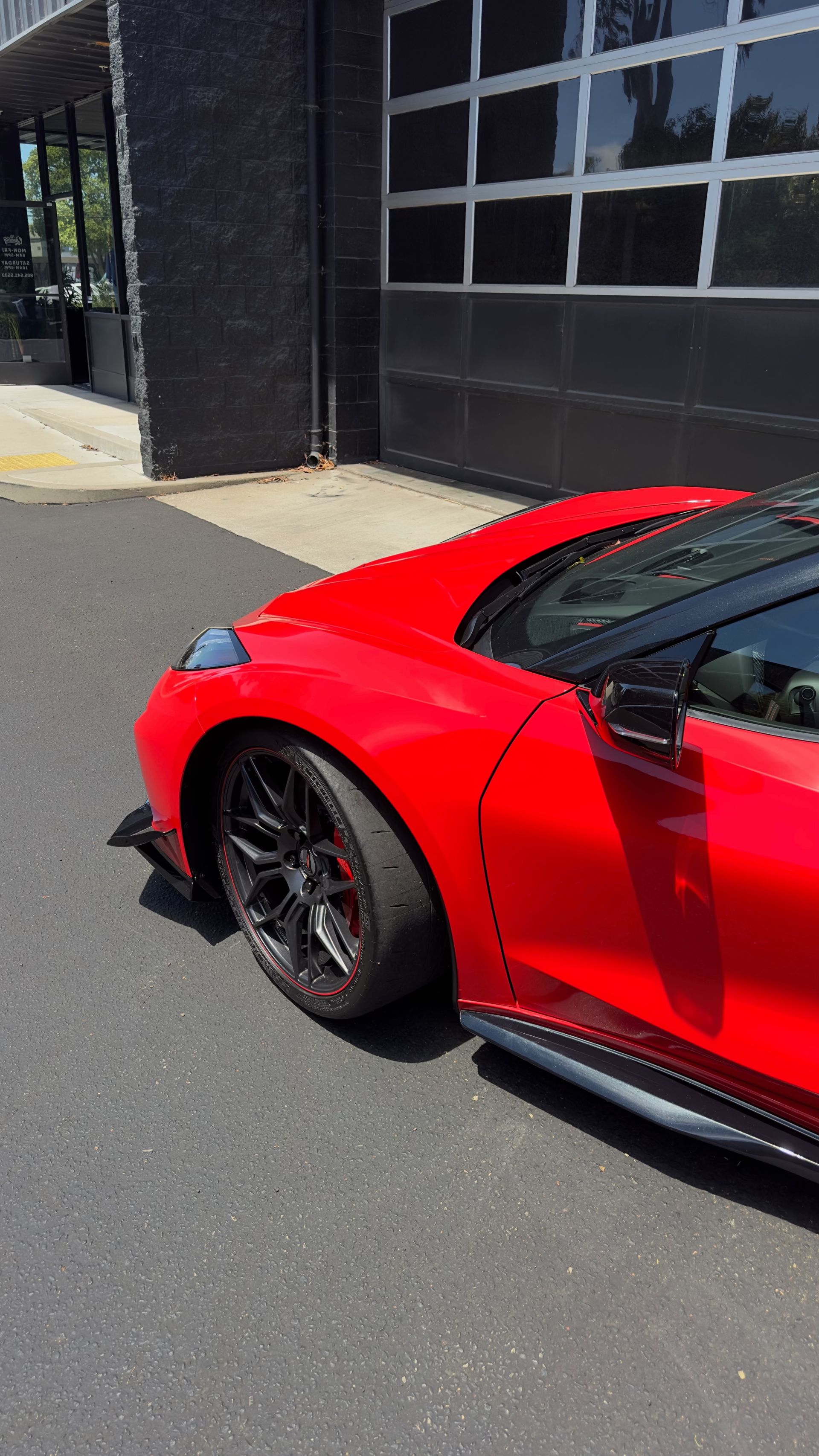 Red sports car parked beside a dark building on a sunny street.