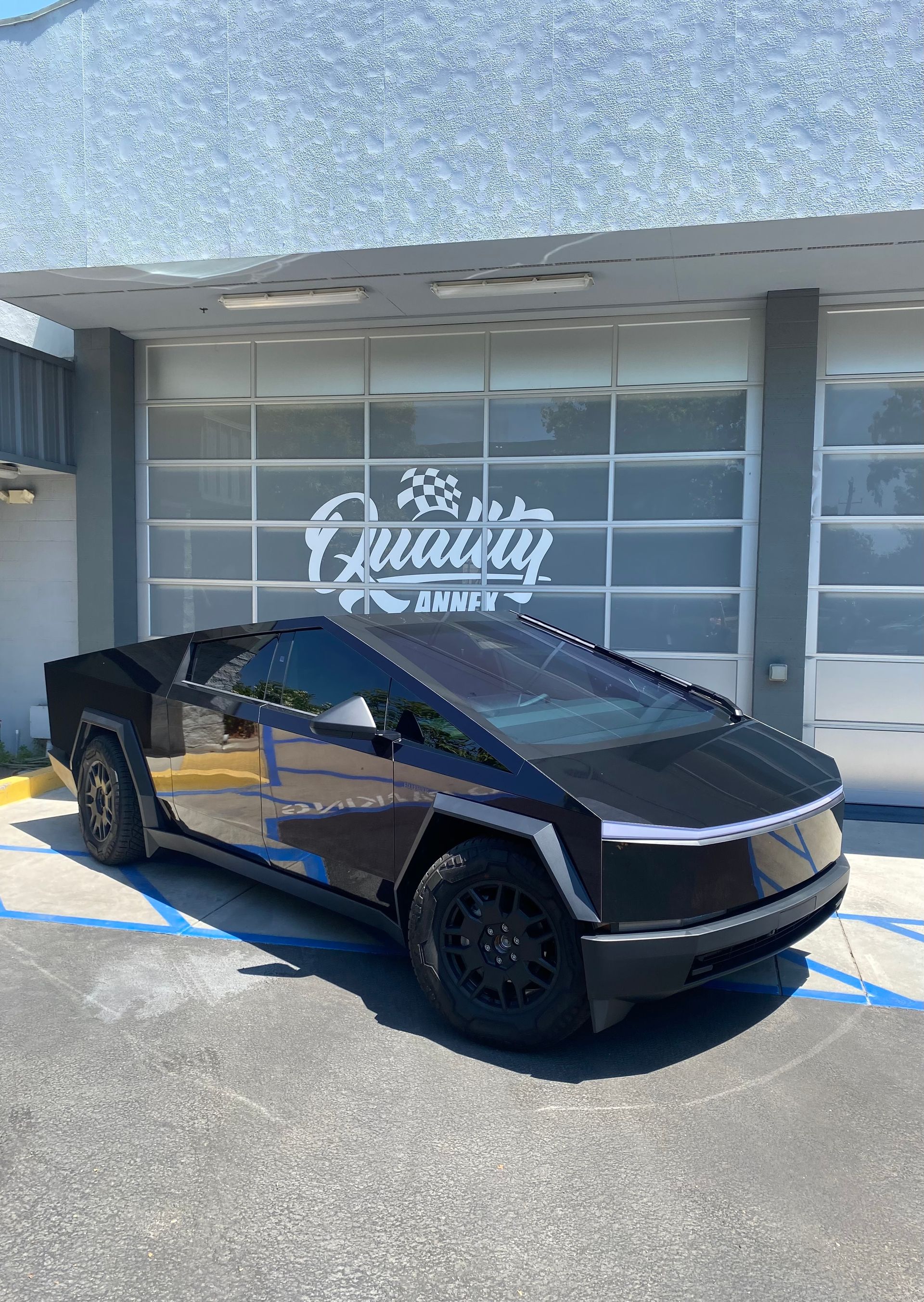 Black Tesla Cybertruck parked outside a dealership with “The Quality” sign on the glass garage doors