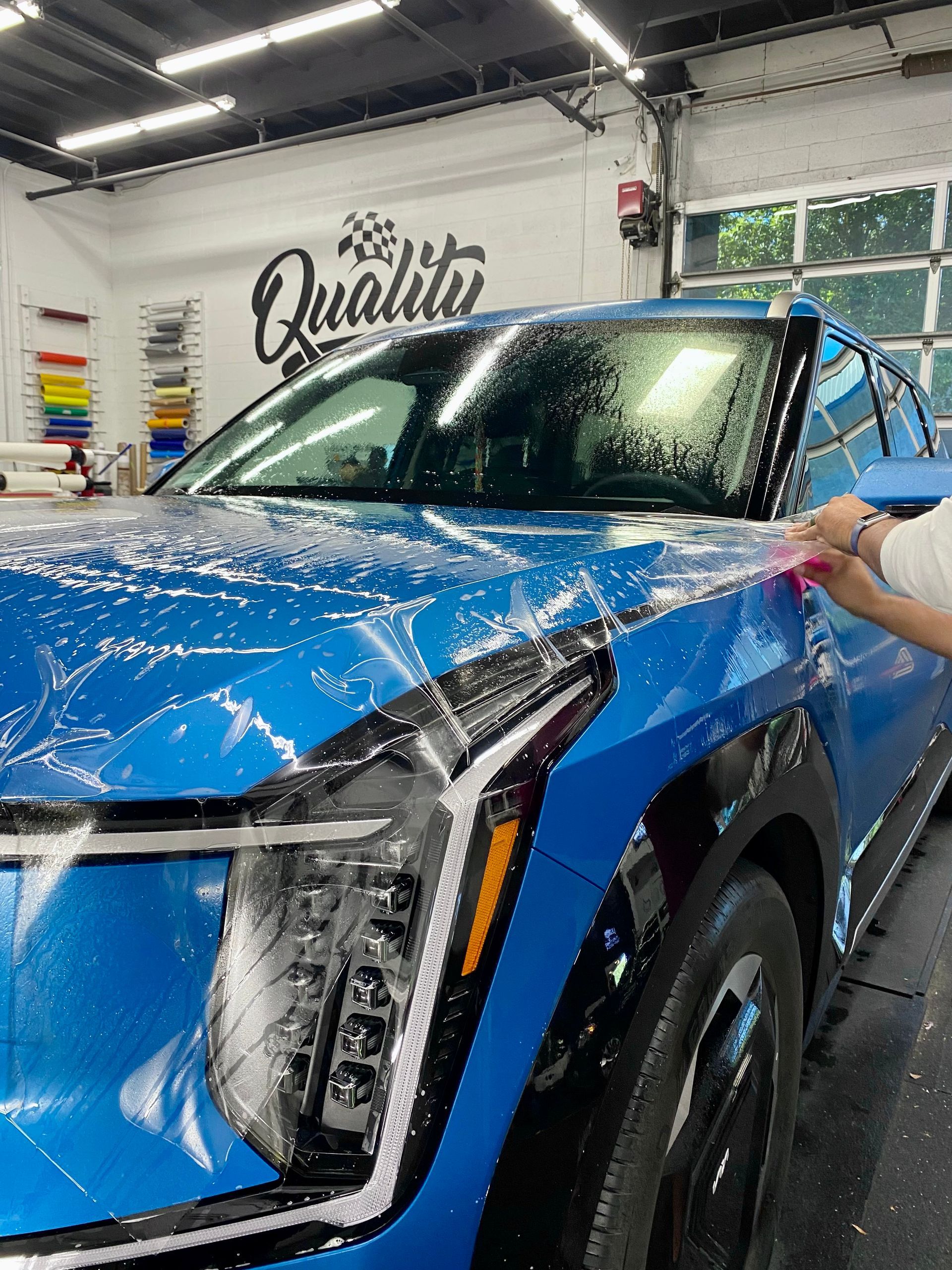 Blue SUV being wrapped in a detailing shop, with a person’s hand smoothing the vinyl on the hood.