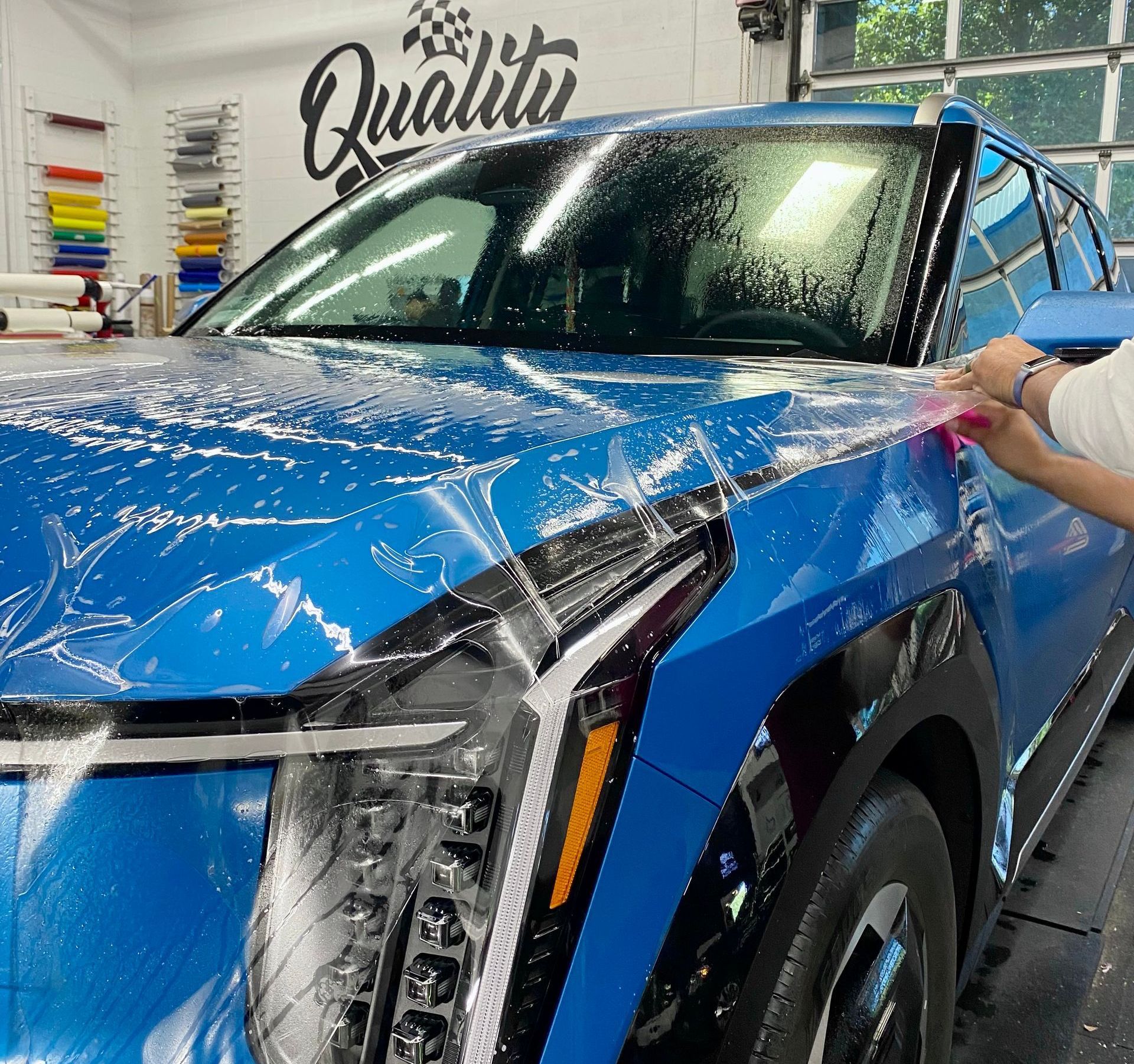 Blue SUV being wrapped in clear protective film in a shop, with a person applying the film to the side.