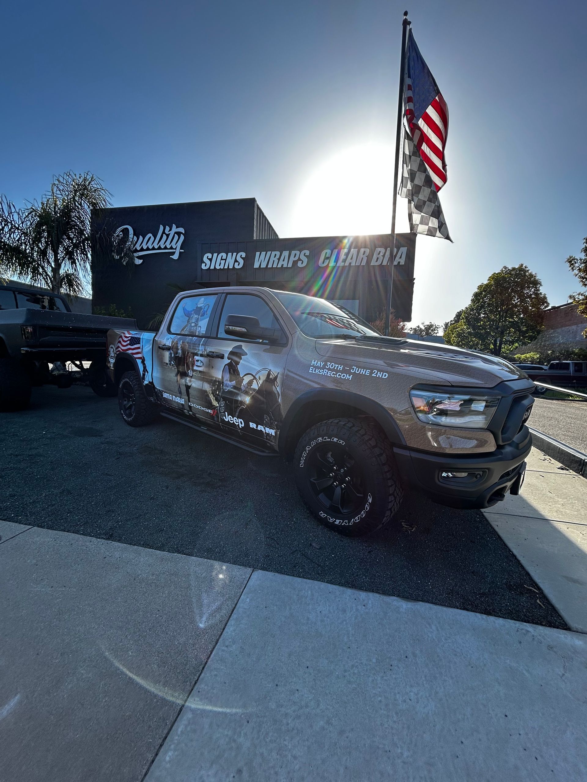 Camouflage pickup truck at an outdoor car show with an American flag and bright sun flare