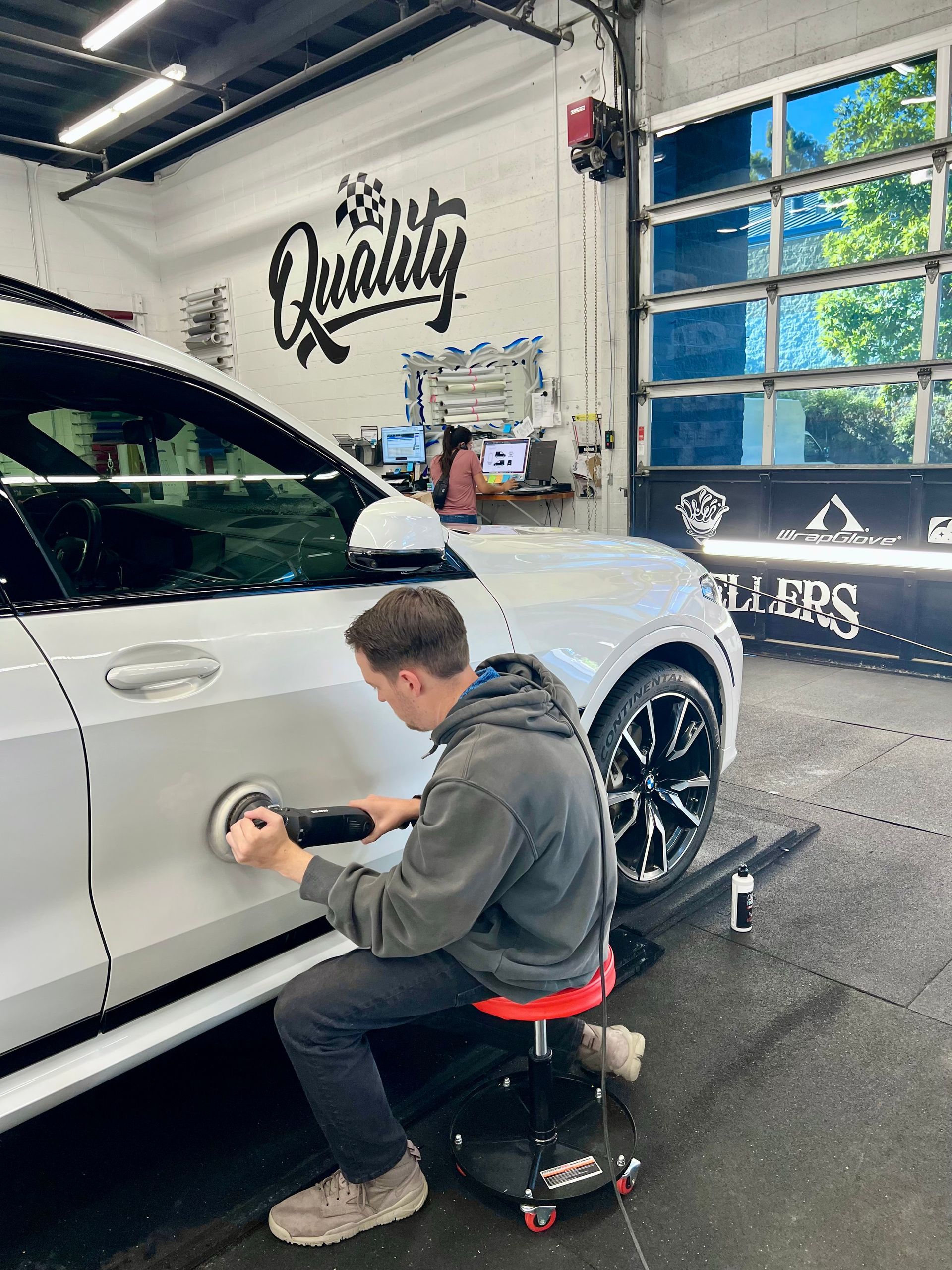 Detailer polishing white SUV door in a car wash bay with “The Quality” sign on the wall