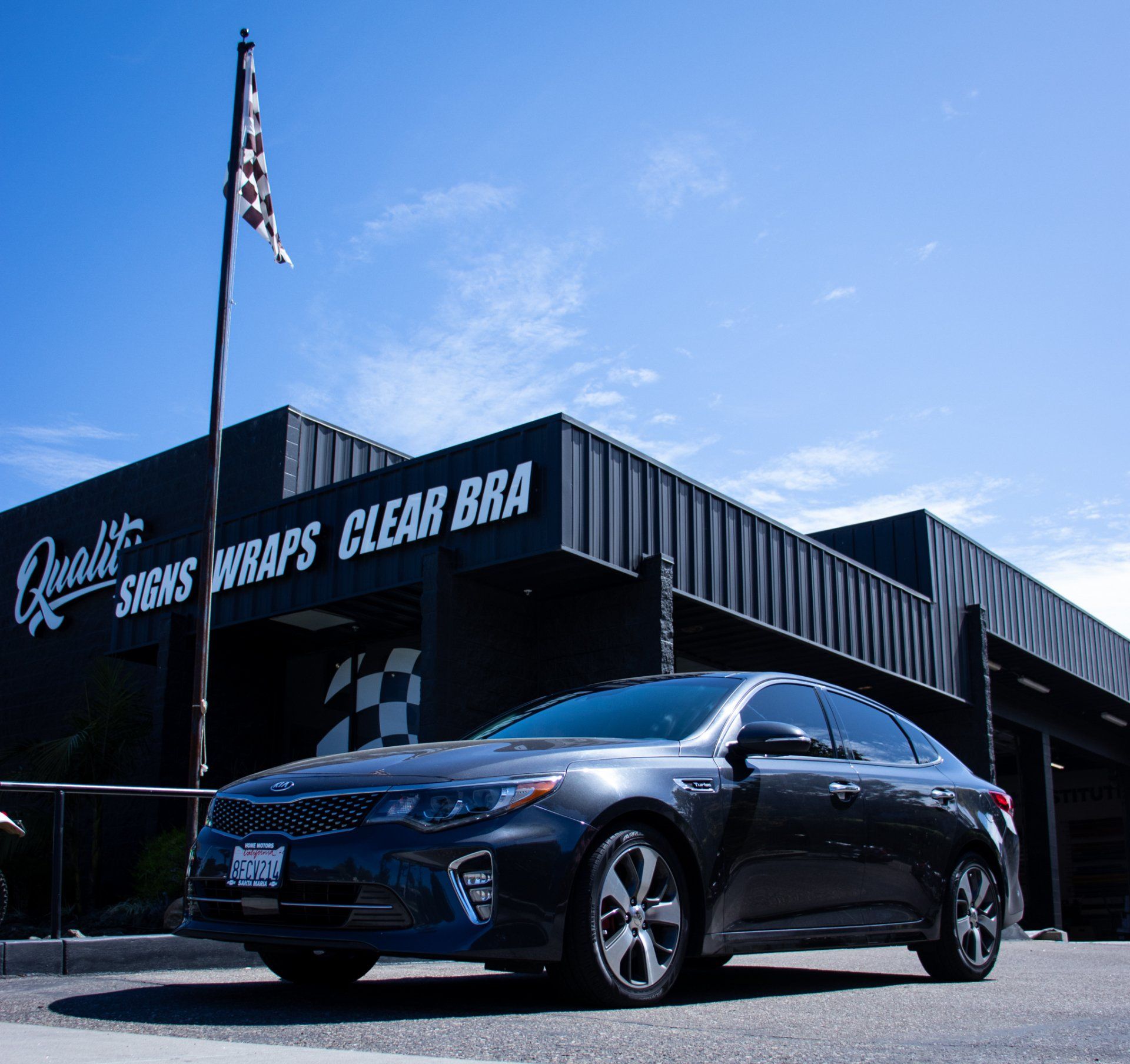 Black sedan parked outside a car dealership under a blue sky.