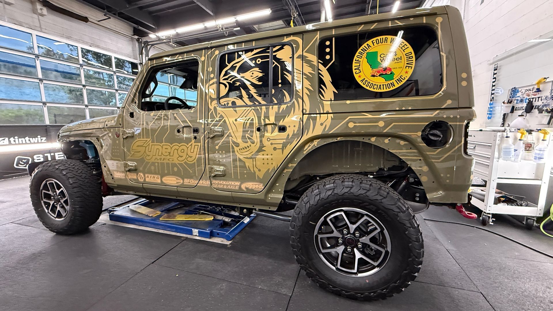 Camouflaged off-road SUV in a garage, raised on a lift with large tires and club-style decals.