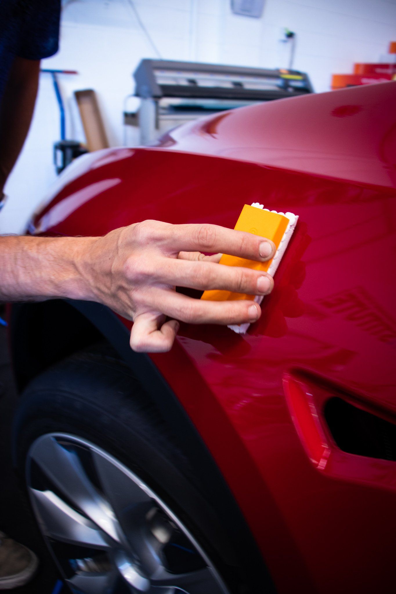 Hand sanding a red car’s body panel with an orange sanding block