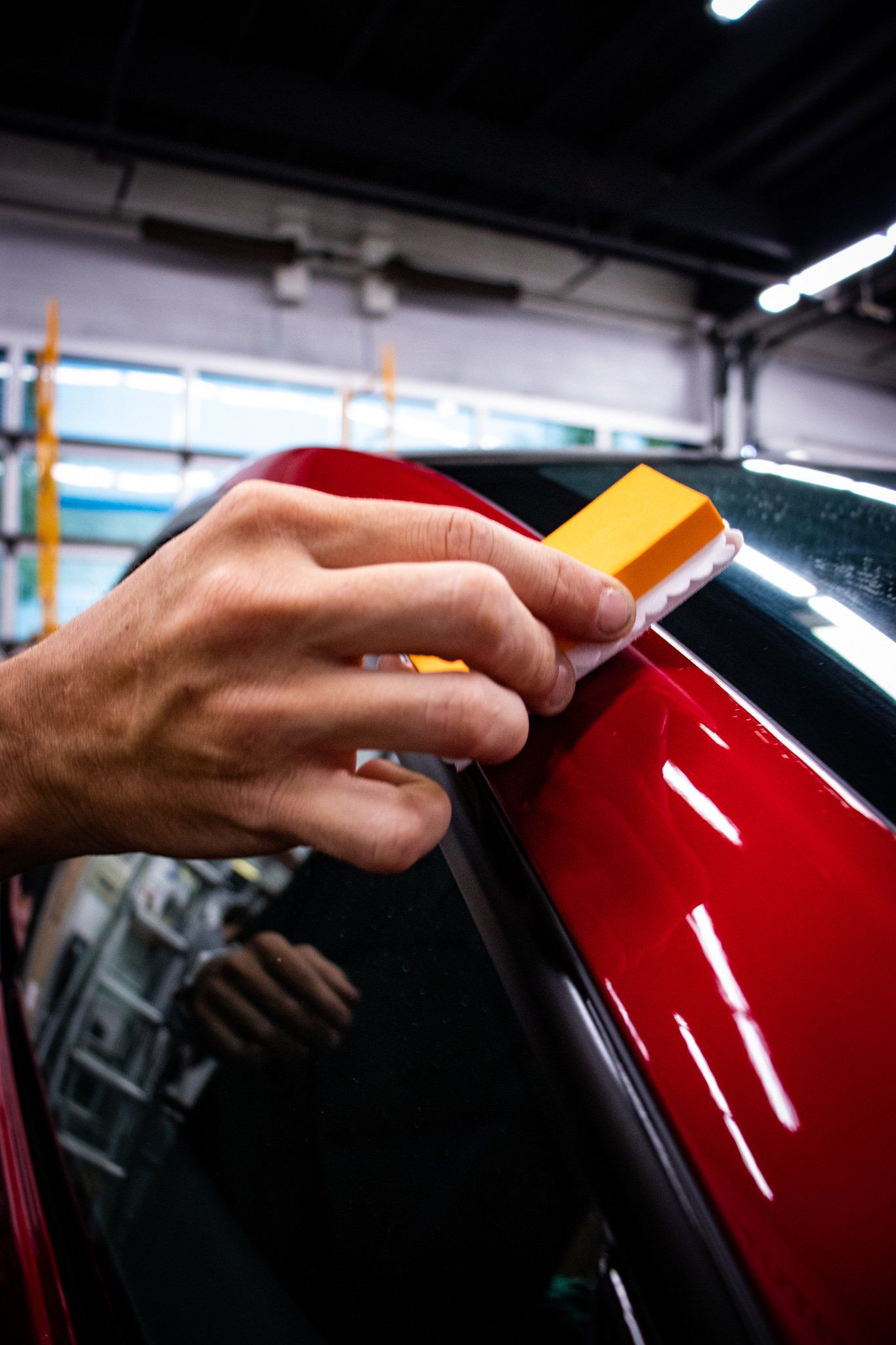 Hand sanding a glossy red car panel with a yellow sanding block in an auto shop