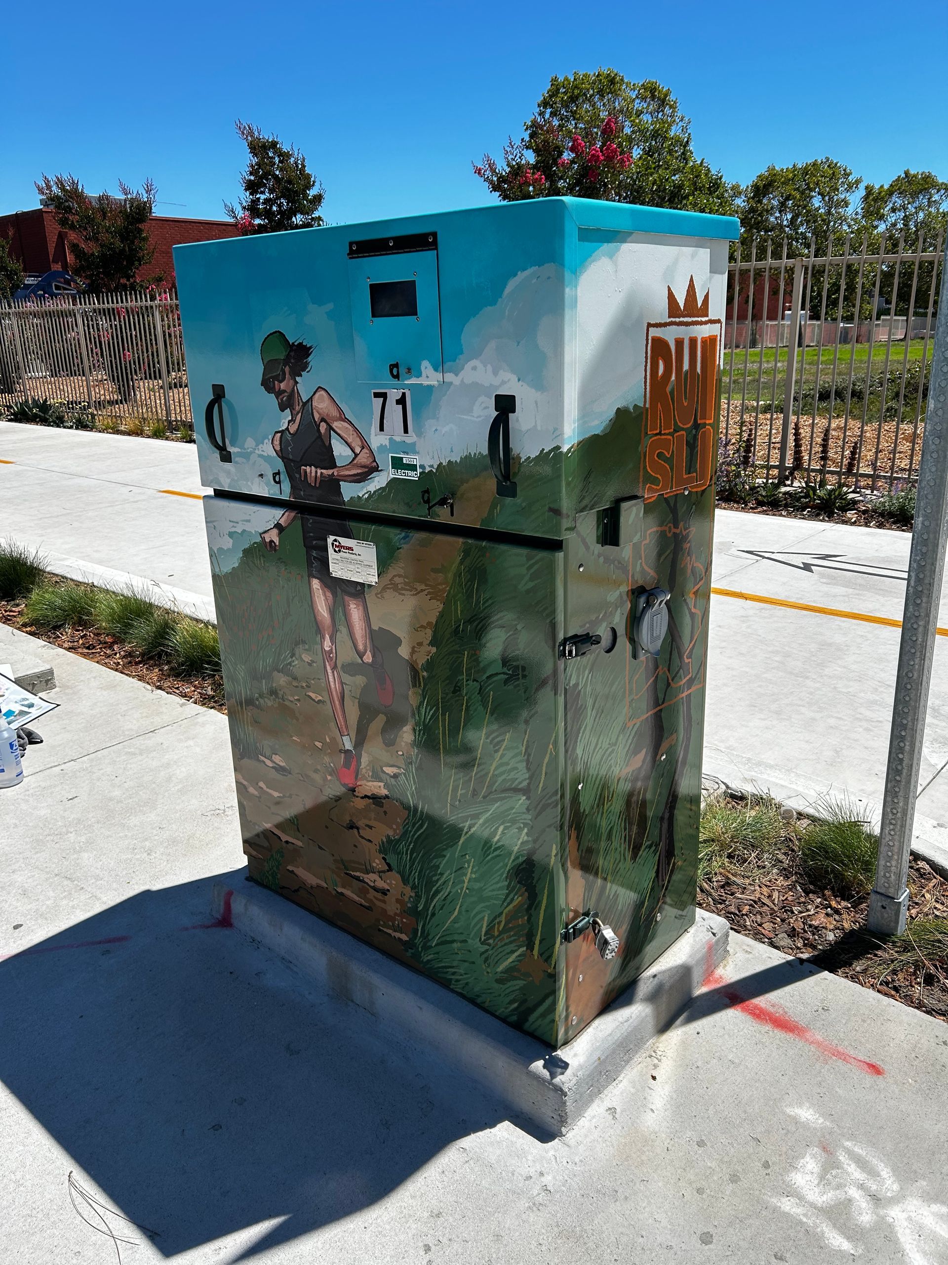 Painted utility box with a mural of a person in a desert scene beside a sidewalk and planter