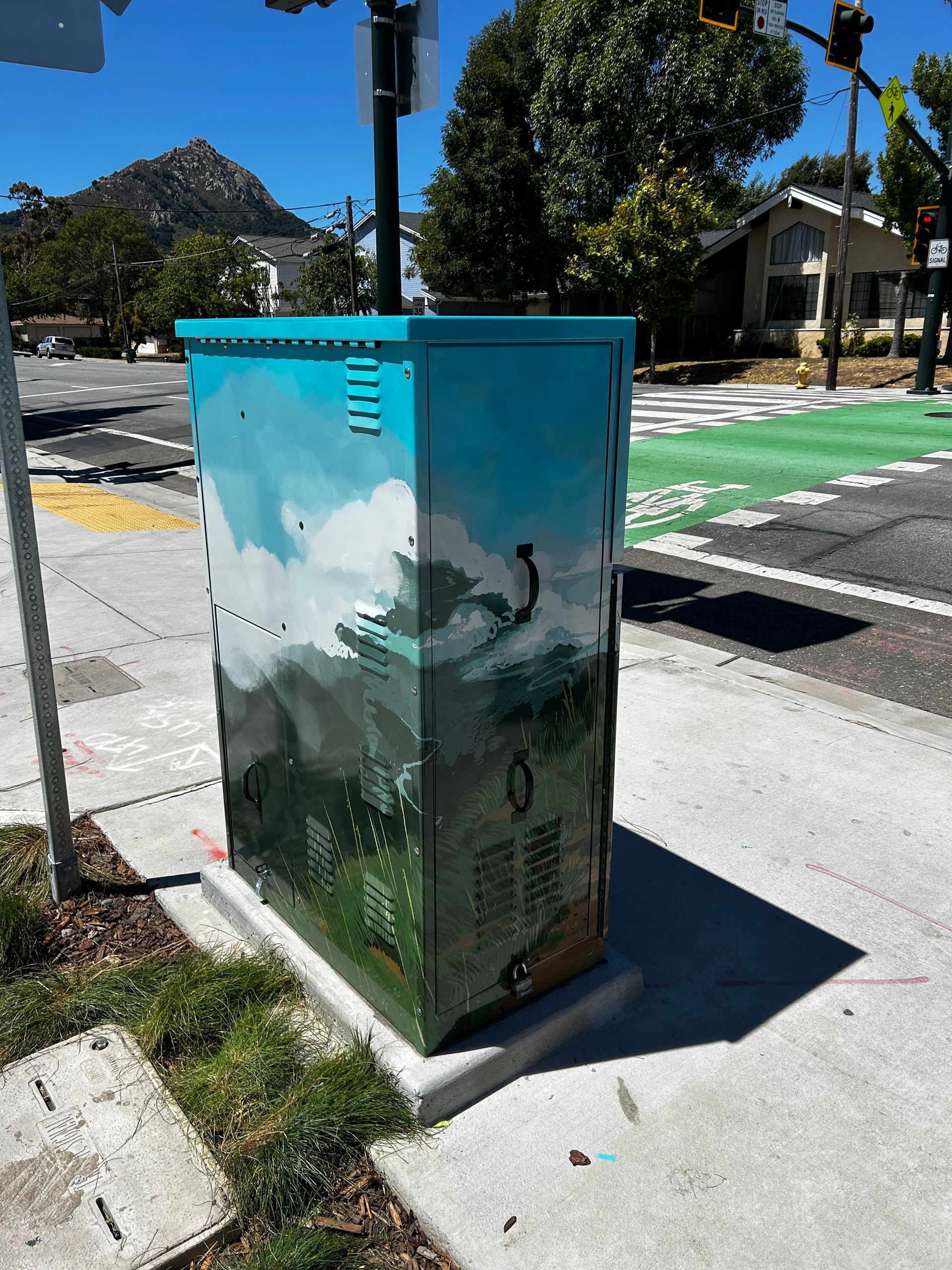 Teal utility box painted with mountain scene beside a sidewalk and crosswalk at an intersection