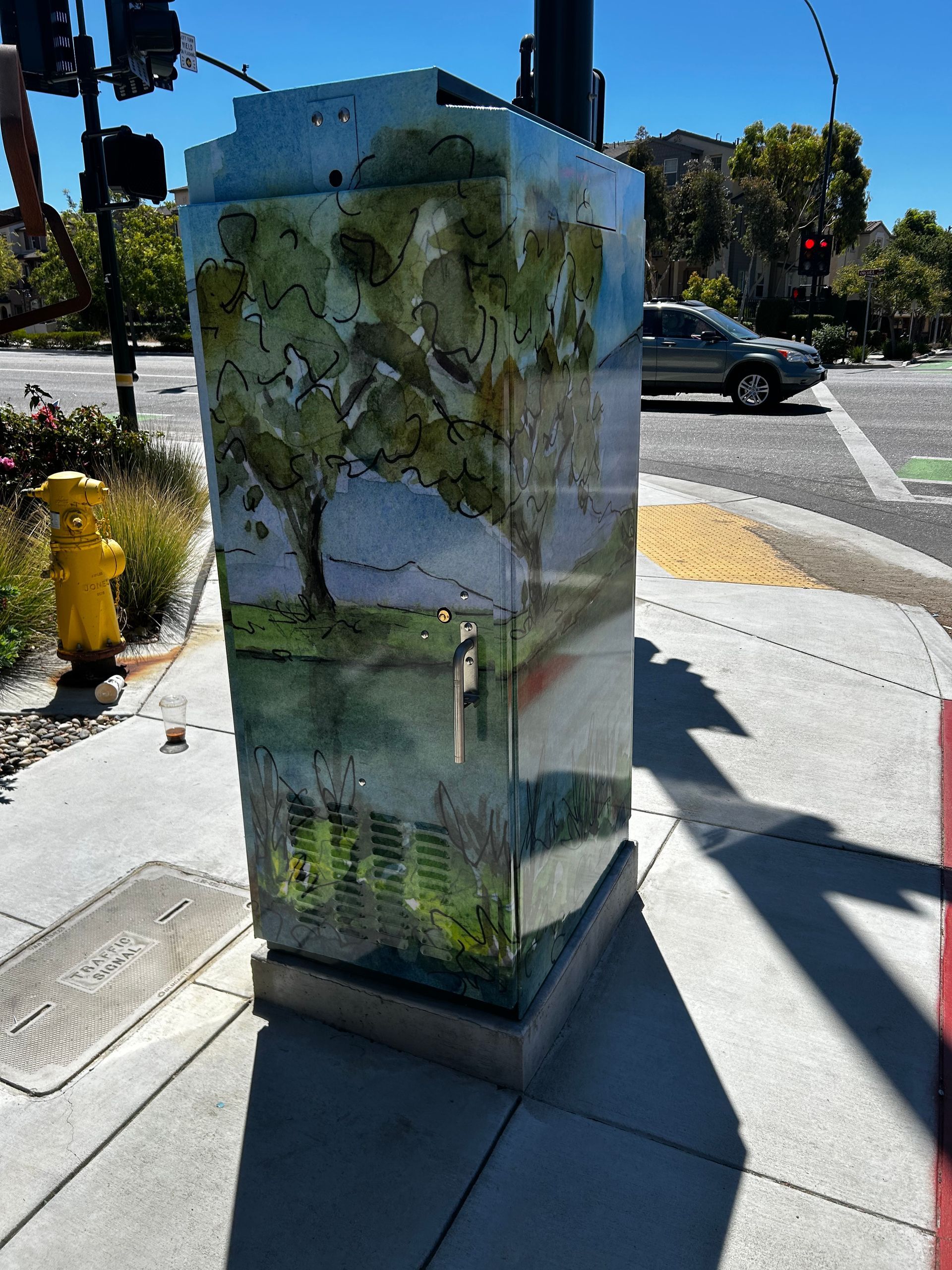 Traffic signal control box wrapped in a green nature mural at a sunny street corner, beside a crosswalk and fire hydrant