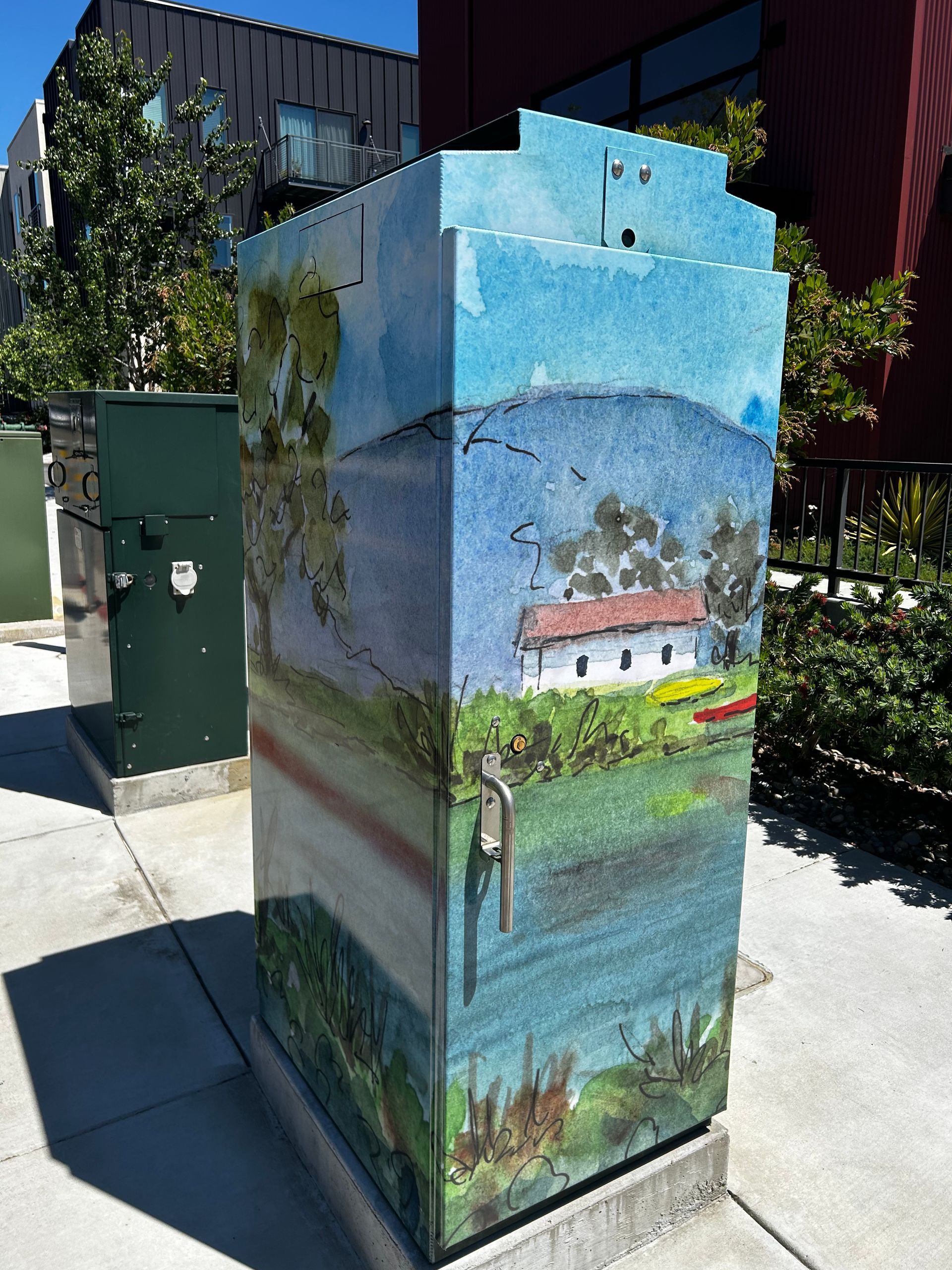 Painted utility box with a landscape mural of a house by water in a sunny outdoor setting