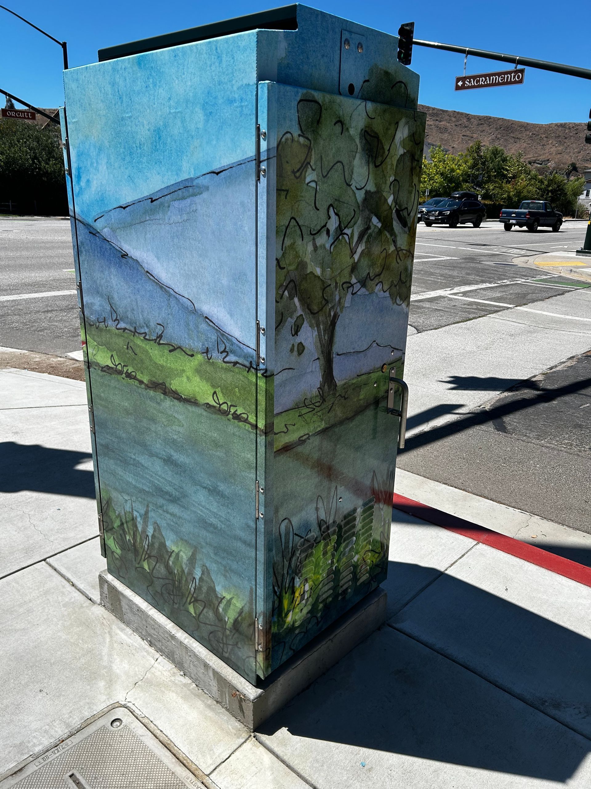 Painted utility box on a sidewalk, decorated with a landscape mural of mountains, water, and greenery.