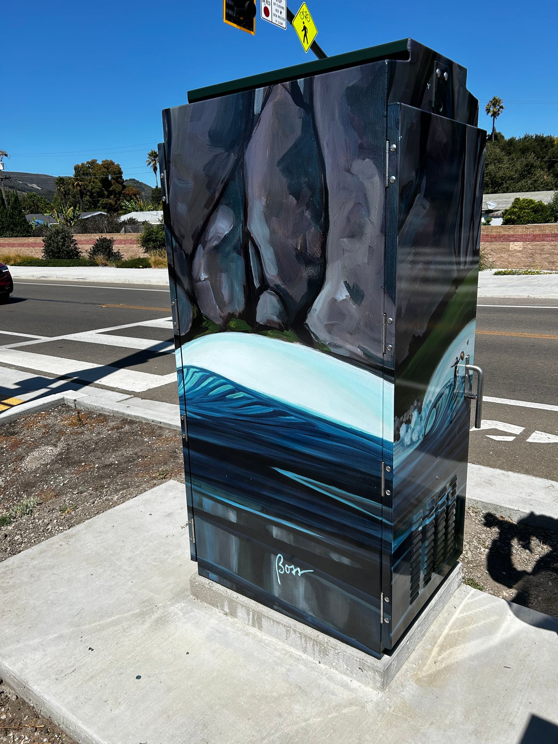 Painted utility box with a blue wave and rocky landscape mural on a sidewalk by the road