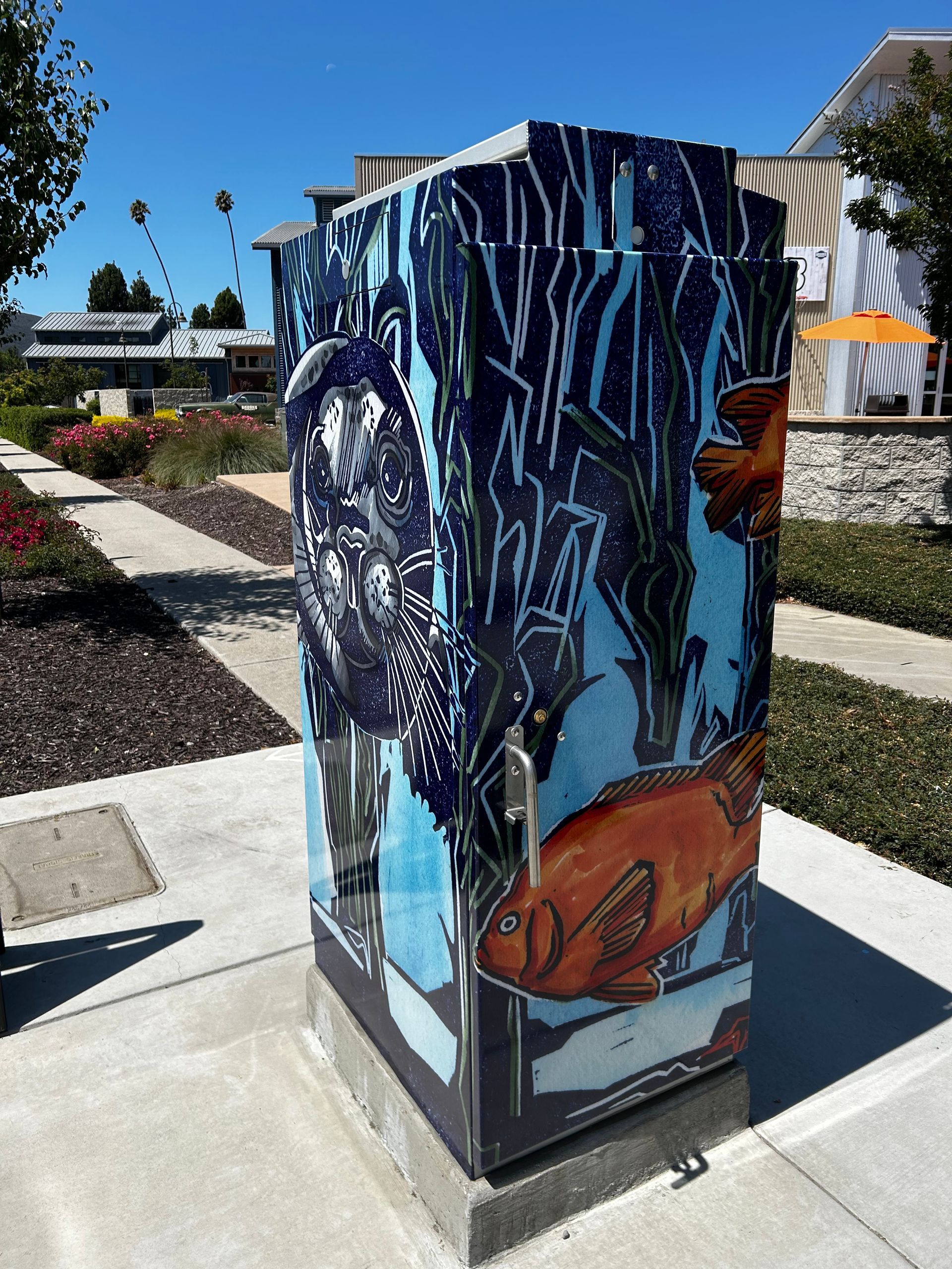 Painted utility box with a blue underwater mural featuring fish and ocean plants on a sidewalk.