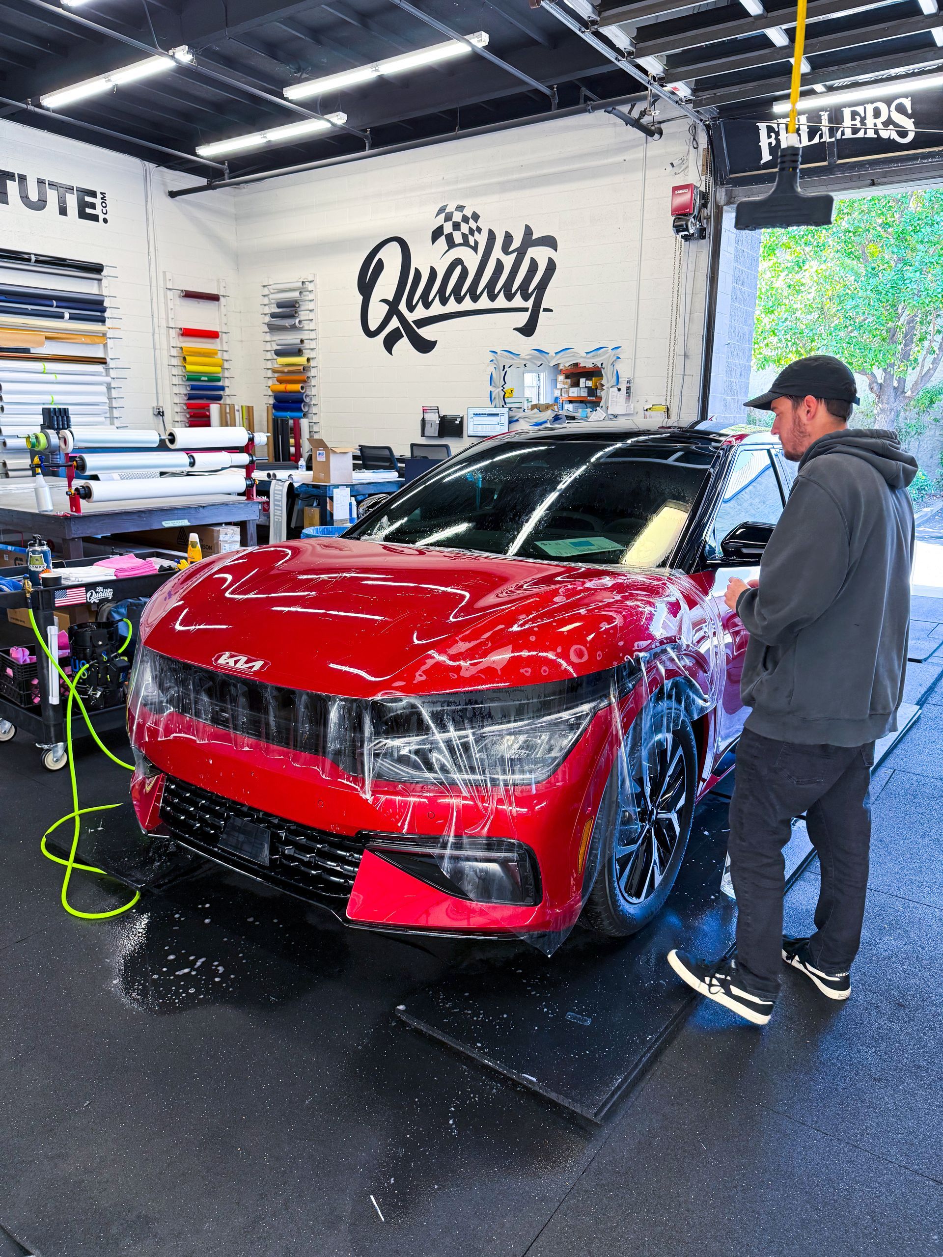Red car being washed indoors by a person in a detailing shop with a “Quality” sign on the wall
