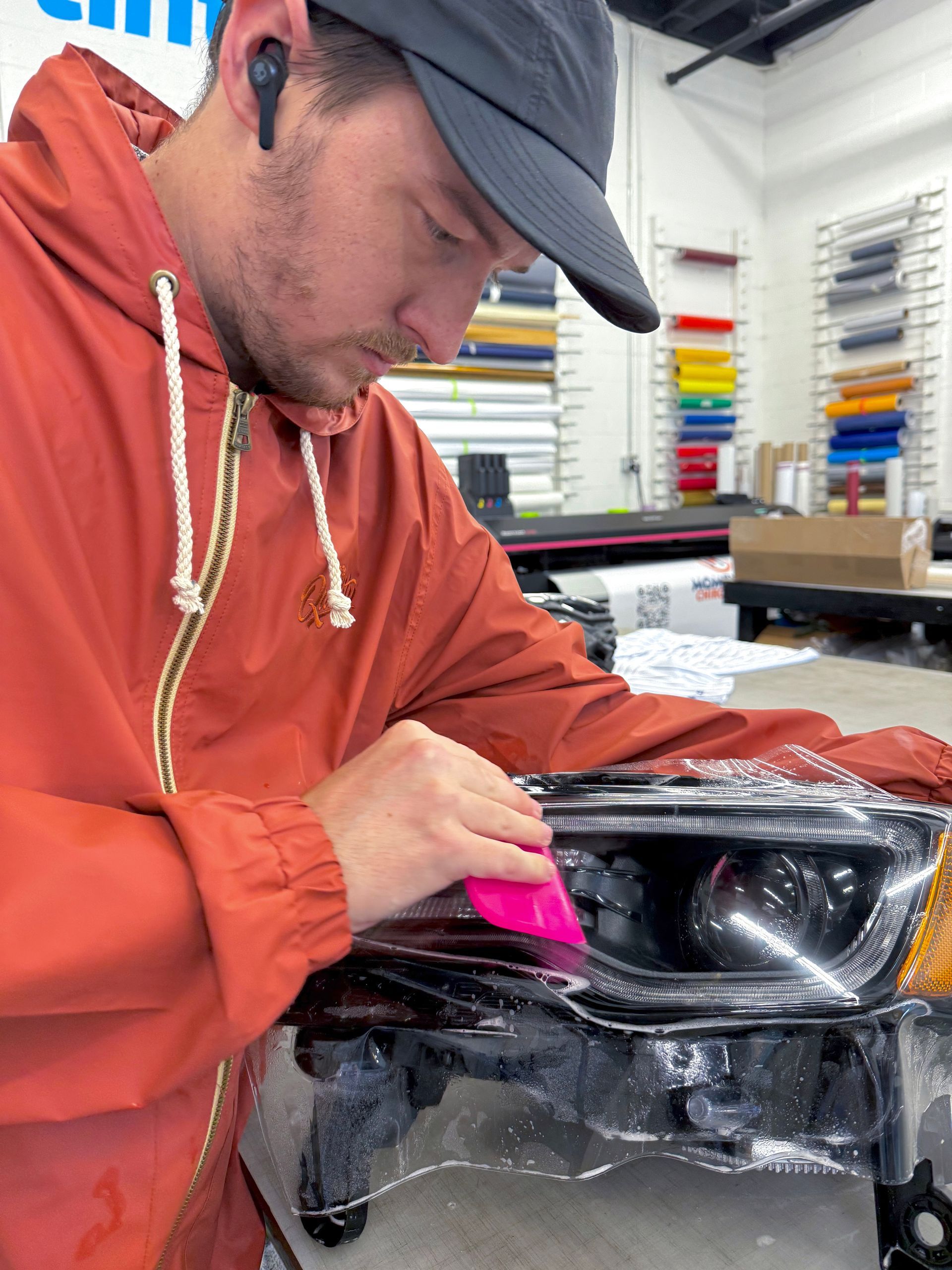 Worker in orange hoodie and cap inspecting a car headlight assembly with pink tape in a workshop