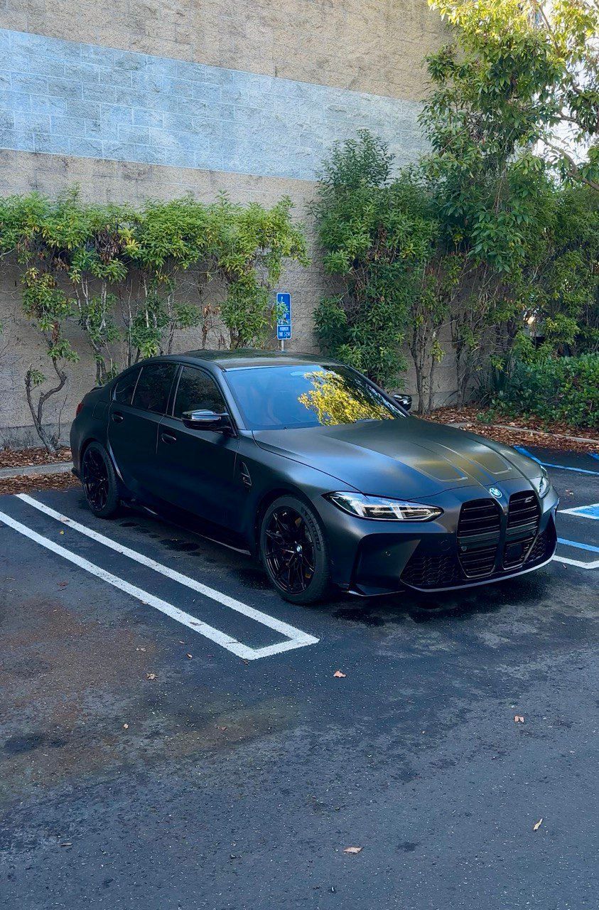 Black sedan parked in a lot beside a wall and greenery, with blue accessible parking markings nearby