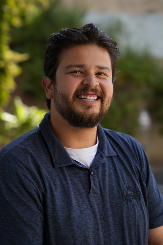 Smiling man in a navy polo shirt standing outdoors with a blurred green background