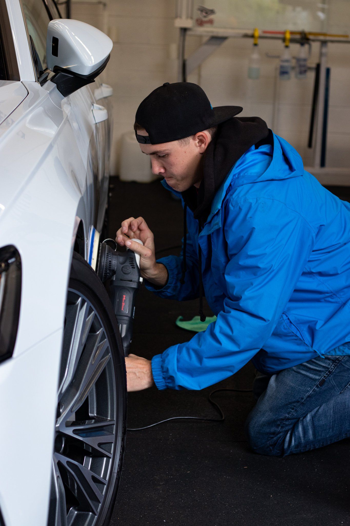 Mechanic in blue jacket kneeling beside a white car, inspecting the wheel with a tool in a garage