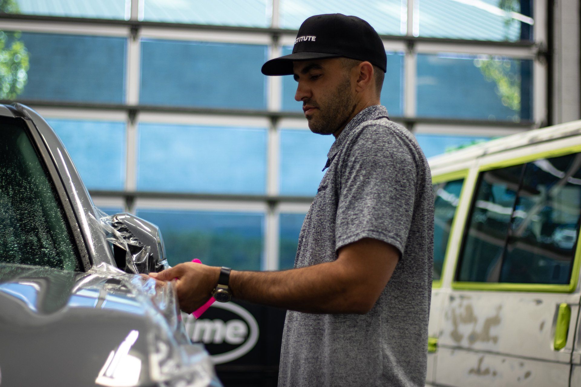 Man in a black cap washing a silver car outdoors beside a white van