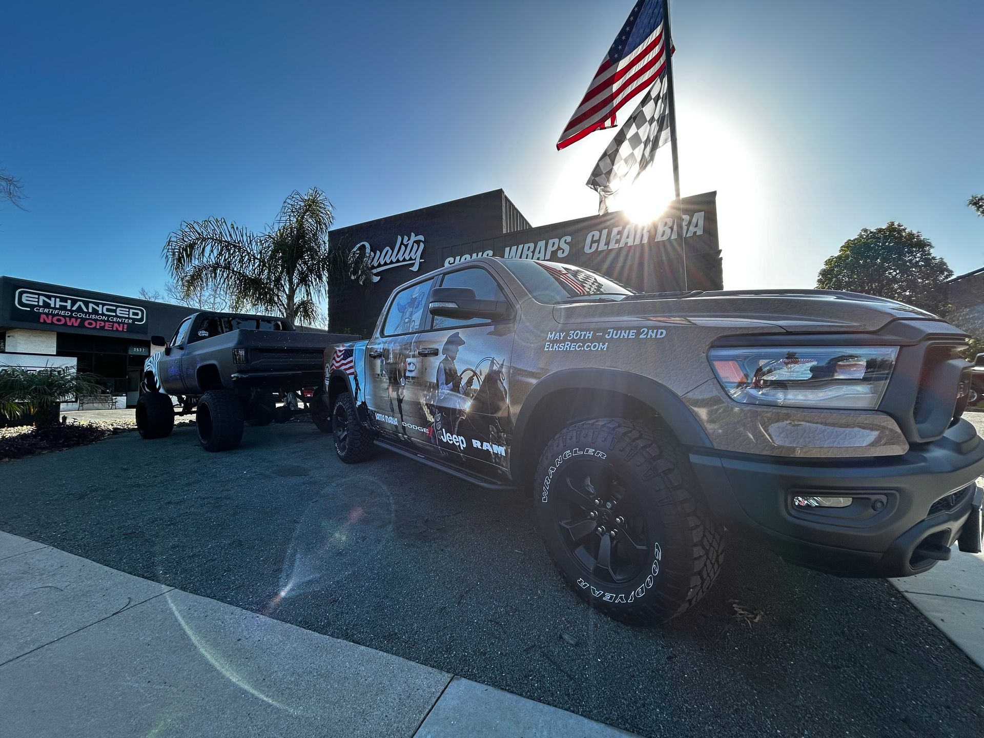 Camouflaged pickup truck with American flag at an outdoor car dealership, sunlit lot.