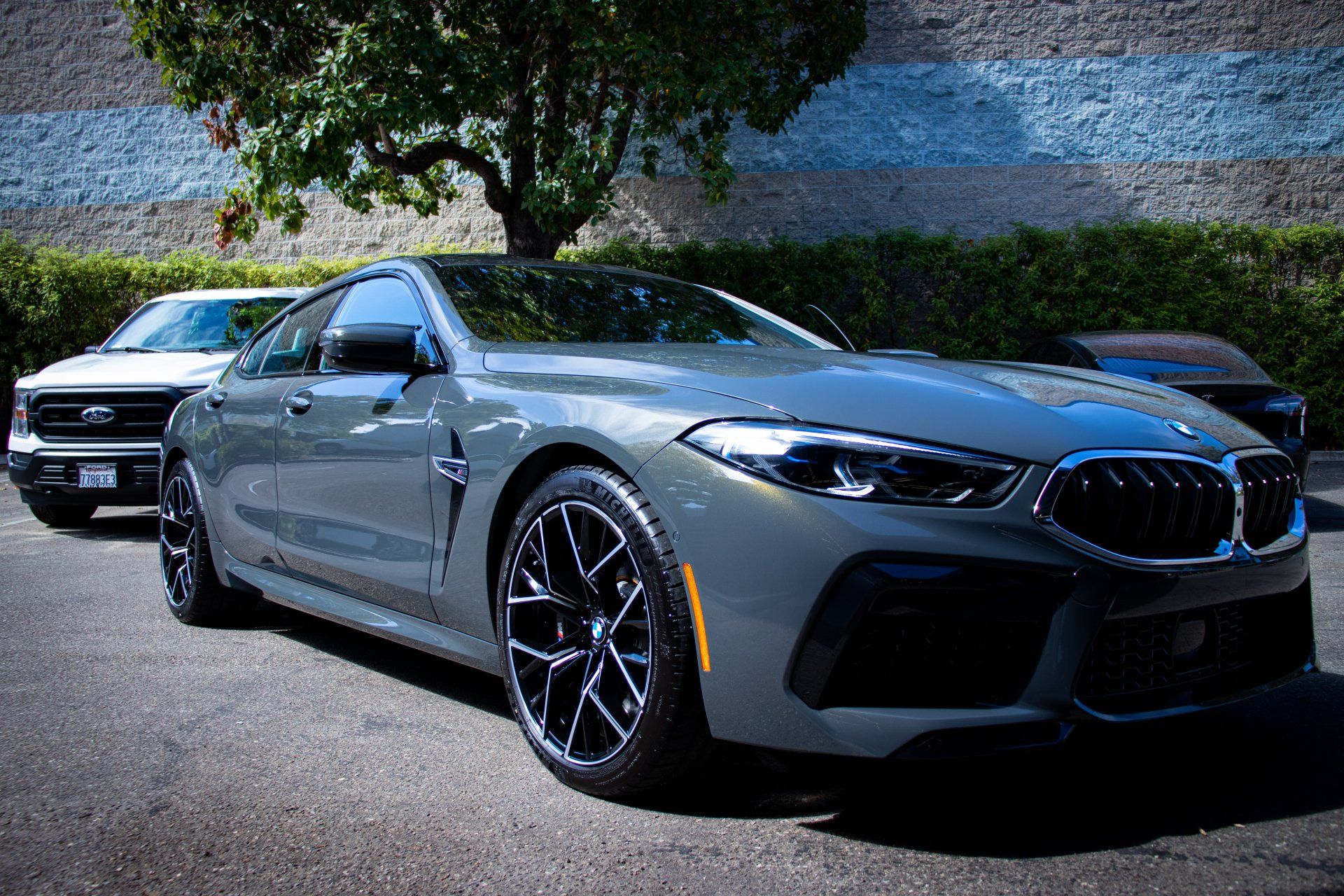 Gray BMW coupe parked outdoors with a black SUV behind it, in front of a stone wall and trees