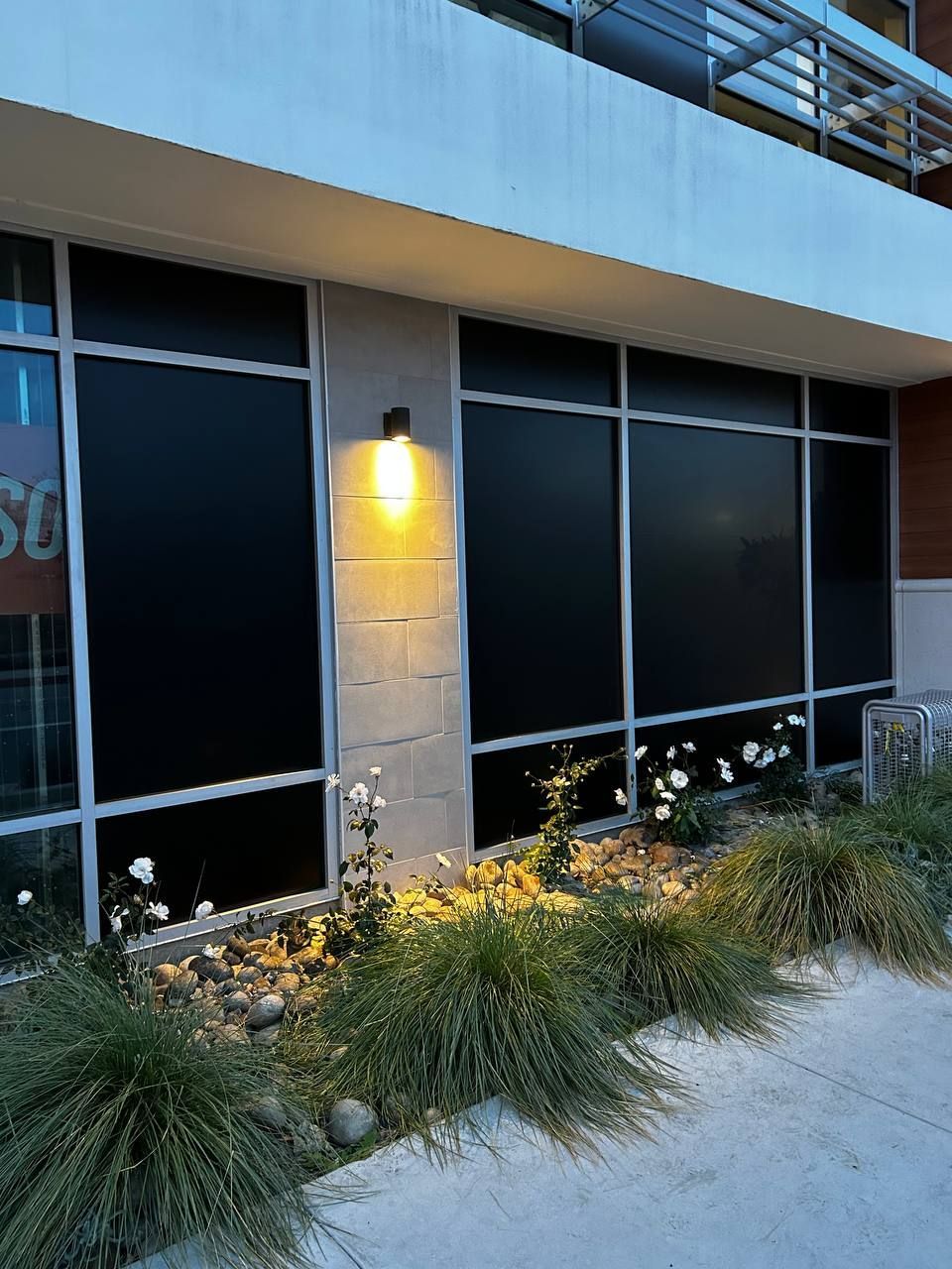 Modern building exterior with black windows, a lit wall sconce, and landscaped grasses by the sidewalk