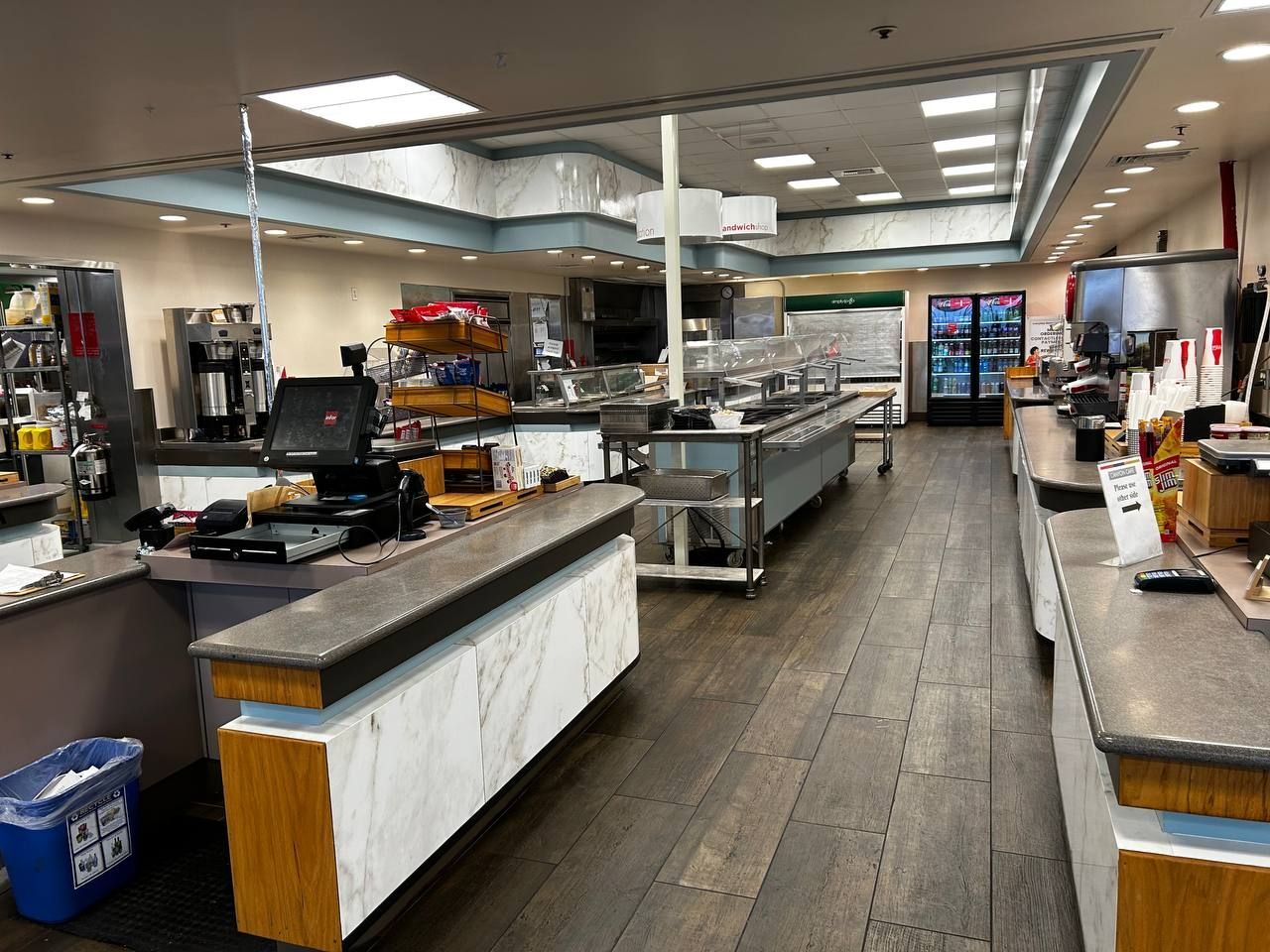 Empty fast-food counter with stainless steel prep tables and a wooden floor inside a cafeteria.