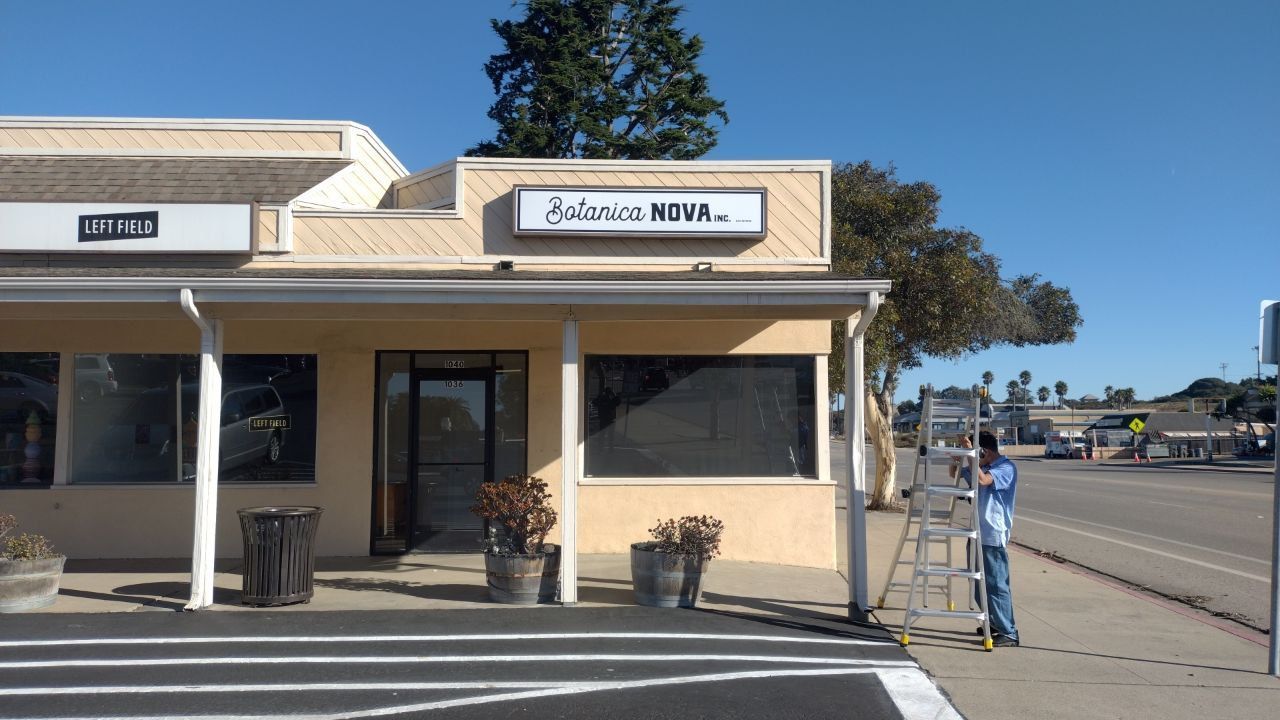 Small beige storefront with a sidewalk sign and parked street under a clear blue sky.