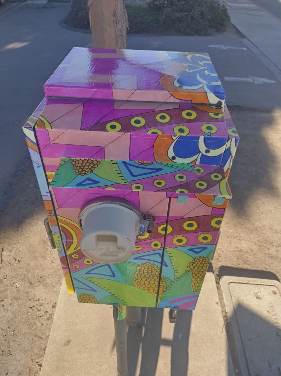Colorful mosaic utility box with a gray circular outlet on a sidewalk near a curb