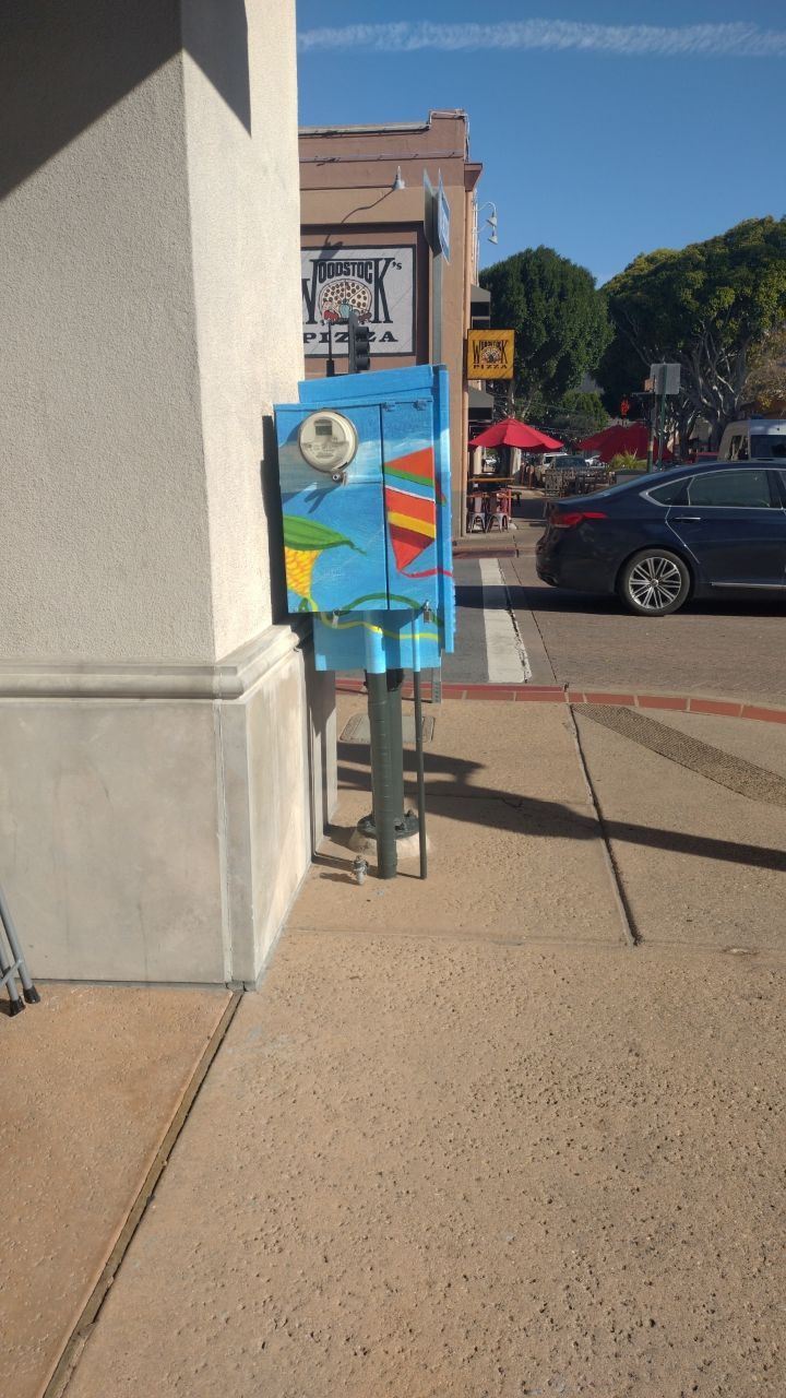 Colorful utility box on a sidewalk near a street, with cars and buildings in the background.