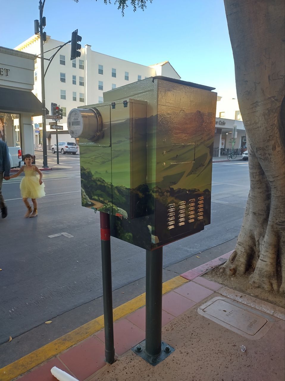 Street-side utility box with mural art on a city sidewalk beside a tree and a child walking nearby