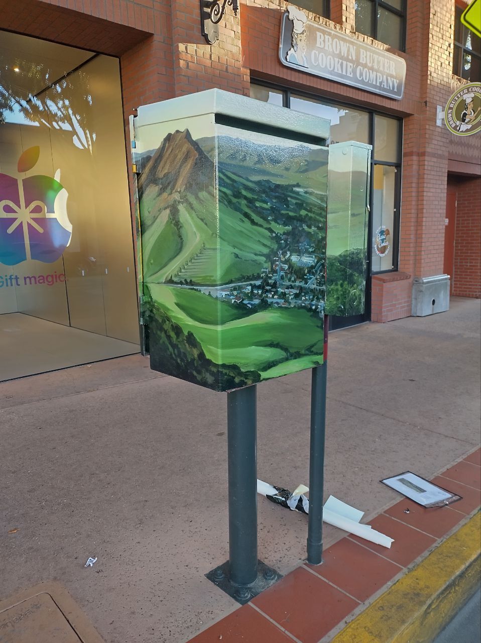 Painted utility box on a sidewalk, showing a green valley landscape mural near storefronts
